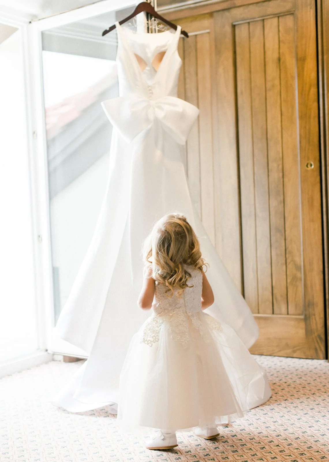 A flower girl looks up at the bride’s wedding dress hanging beside a wooden door.