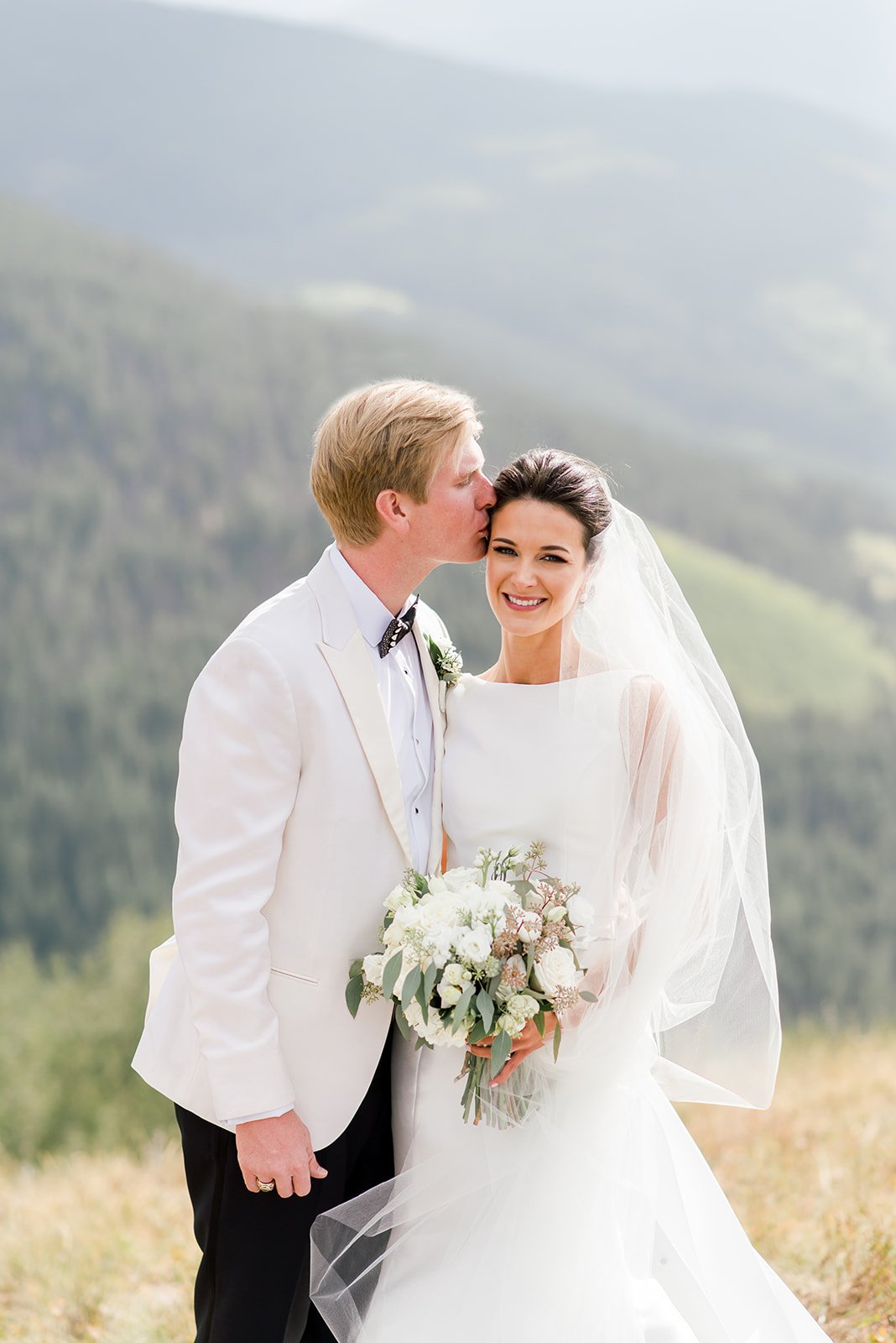 Newlyweds in black-tie attire posing with a white bouquet against the Vail mountains.