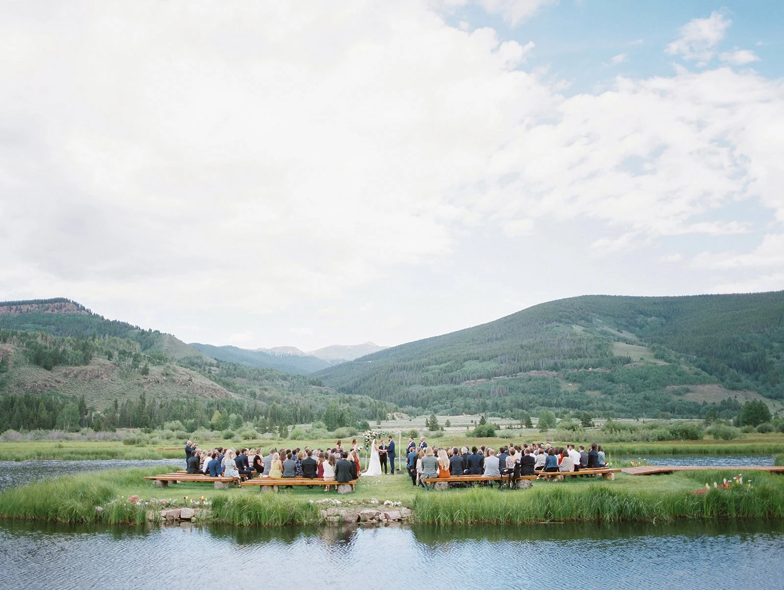 A lakeside wedding ceremony at Camp Hale outside Vail set against rolling Colorado mountain views, with guests seated outdoors for a scenic destination-style celebration. Photo by Sarah Porter Photography.
