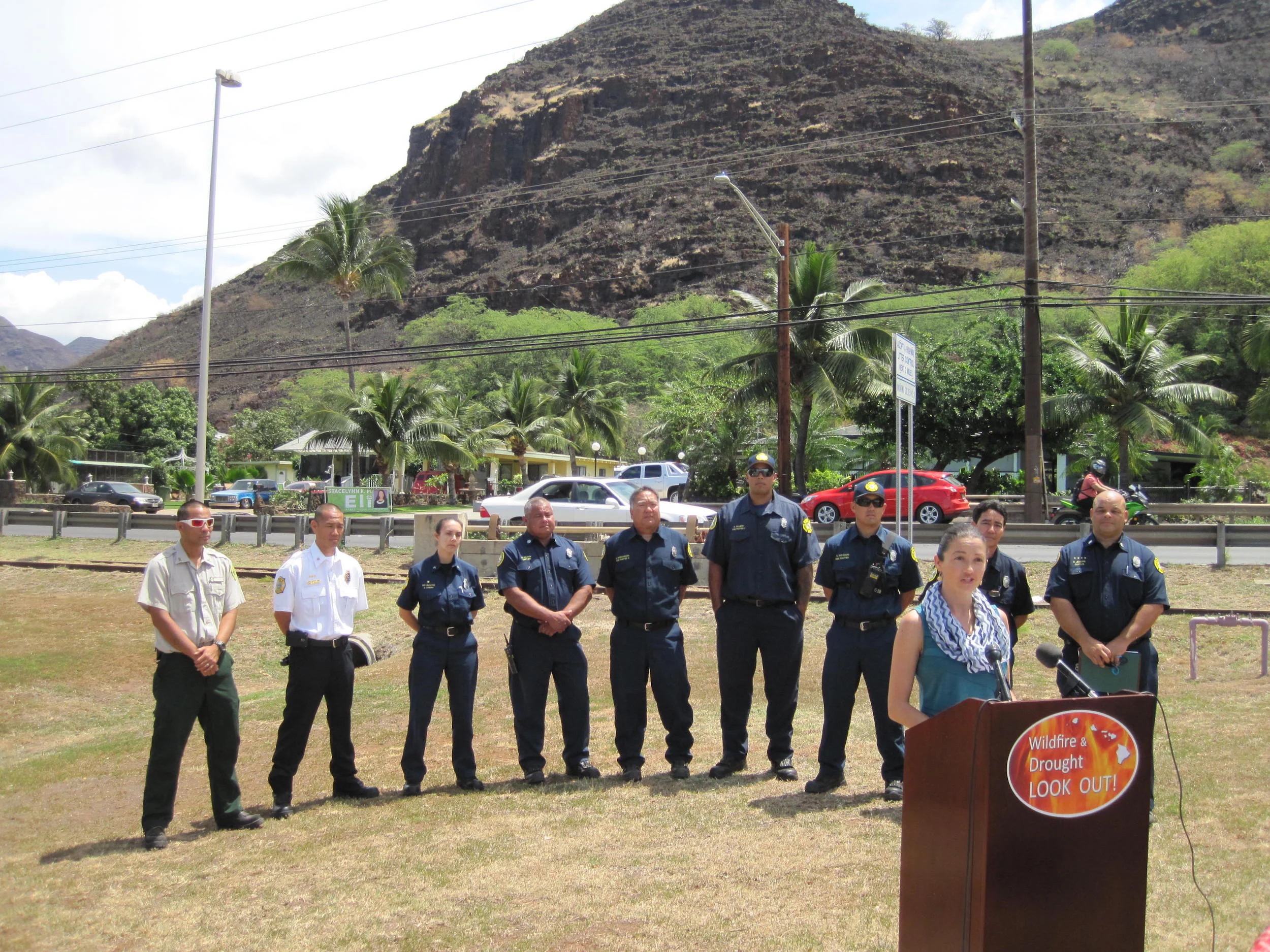 Elizabeth Pickett (HWMO) speaks to media with Oahu firefighters standing behind her. In the background: the charred landscape of Nanakuli from the recent fire.