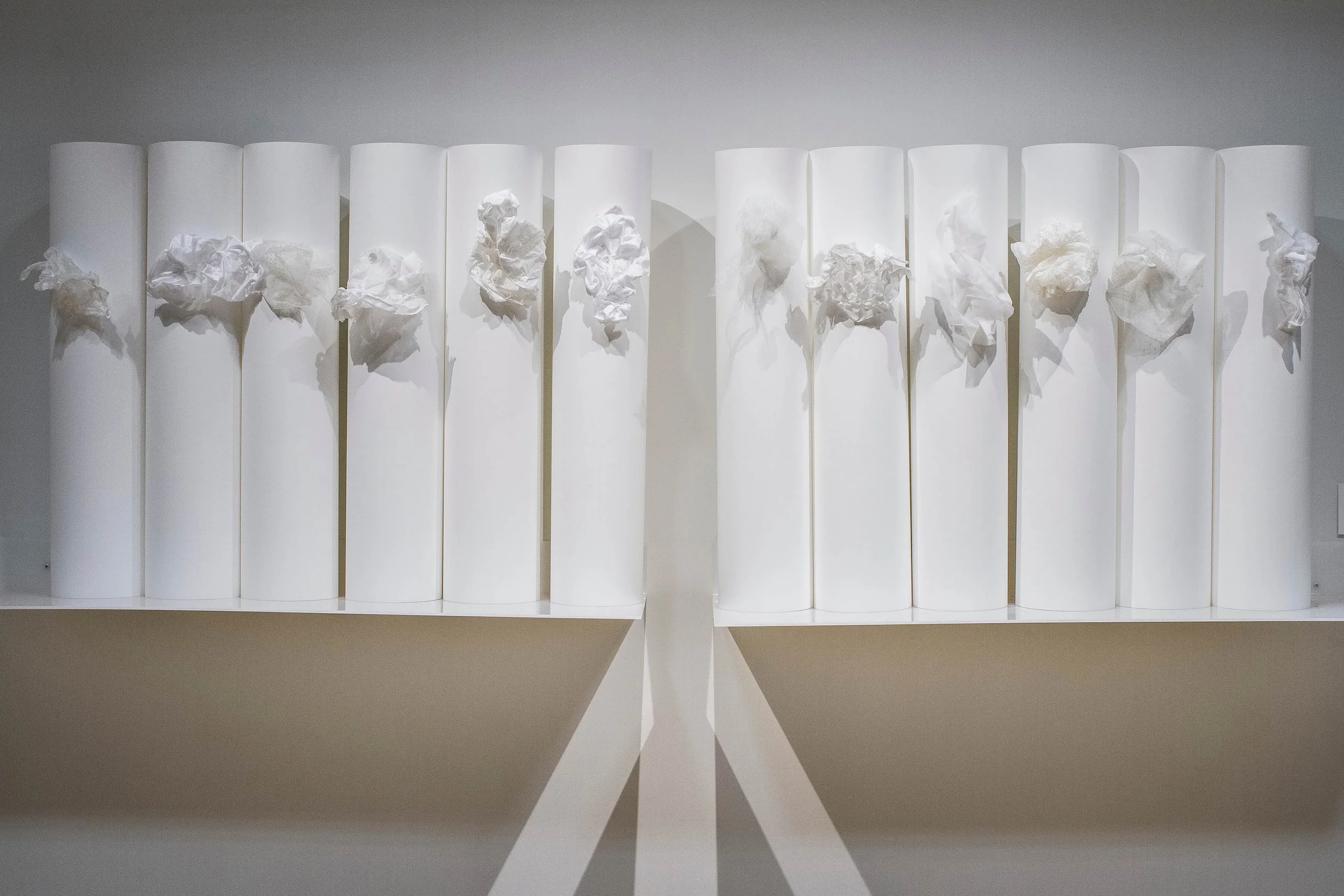 [Installation] UNDERWRITTEN CLOUD DRAWINGS:  Shaped (methylcellulose) cottons and silks buttoned to rag paper (22” x 30”/55.9 x 76.2cm). Paper buttoned up in the back to form cylinders. 12 cylinders on shaped acrylic shelf8 ft (2.4 meters), 2012