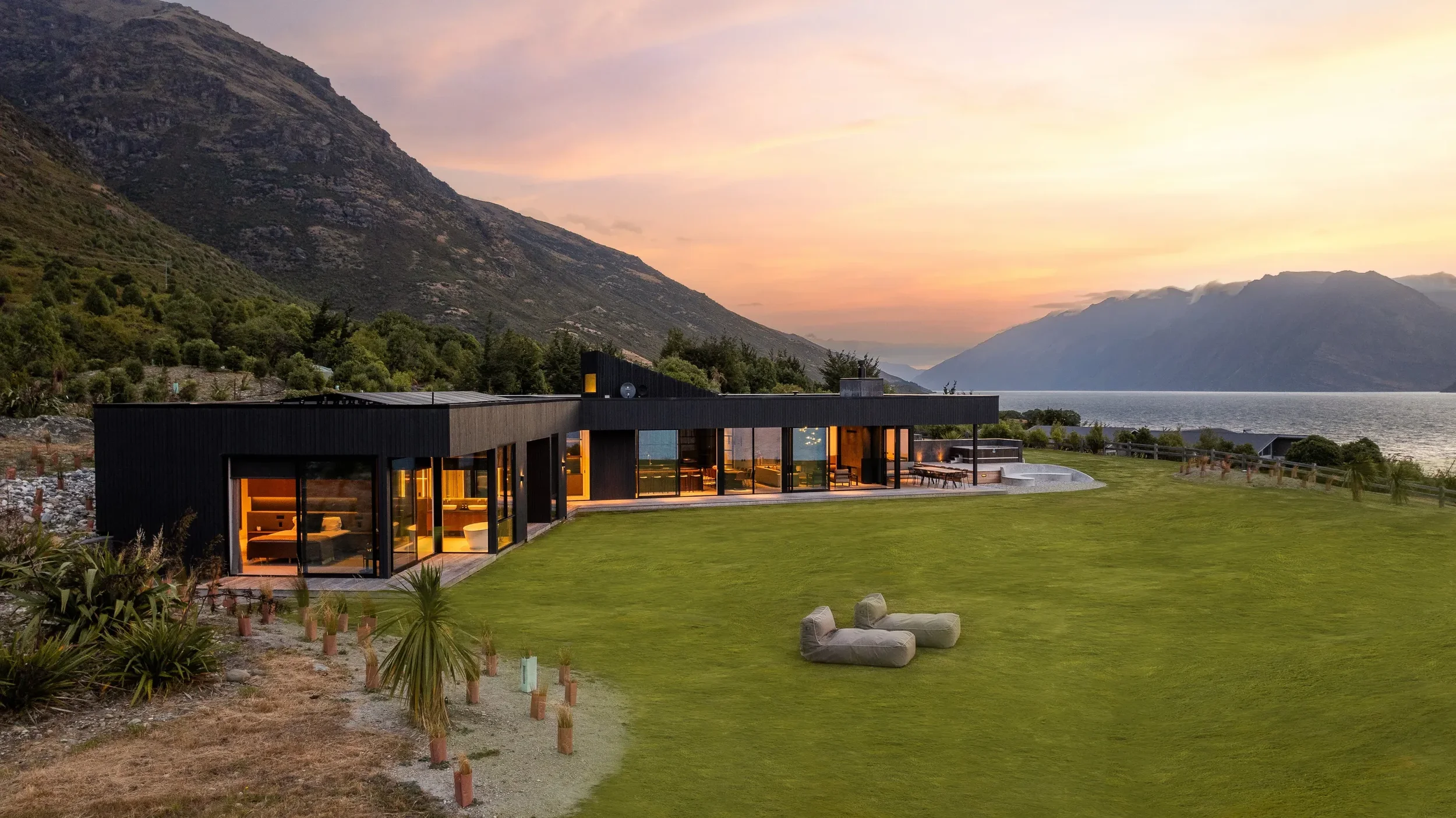 Modern black house with large glass windows overlooking a green lawn and water, mountains in the background during sunset.