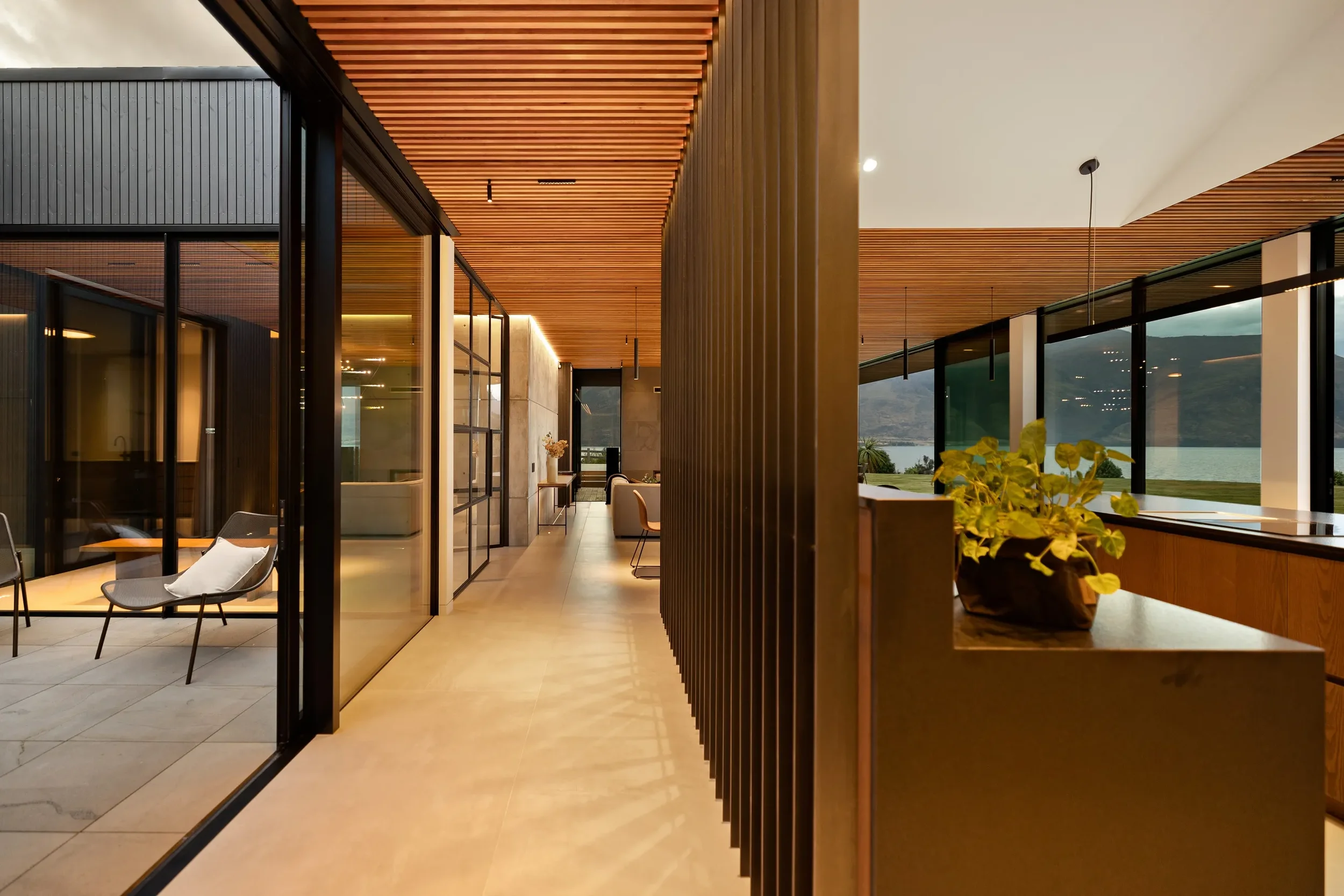 Modern interior with glass walls, wooden ceiling and partitions, outdoor views, chairs, a potted plant, and natural lighting.