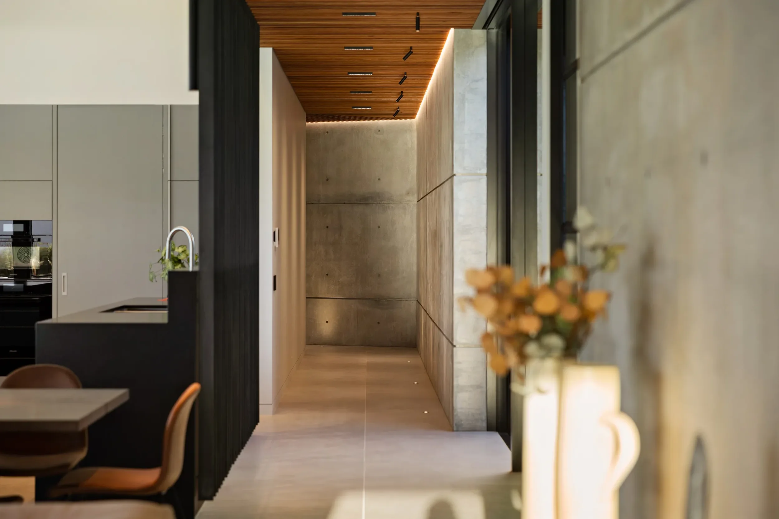 Modern interior hallway with concrete walls, wooden ceiling with recessed lighting, and a hallway leading to a plain wall. A blurred vase with flowers is visible in the foreground on the right.