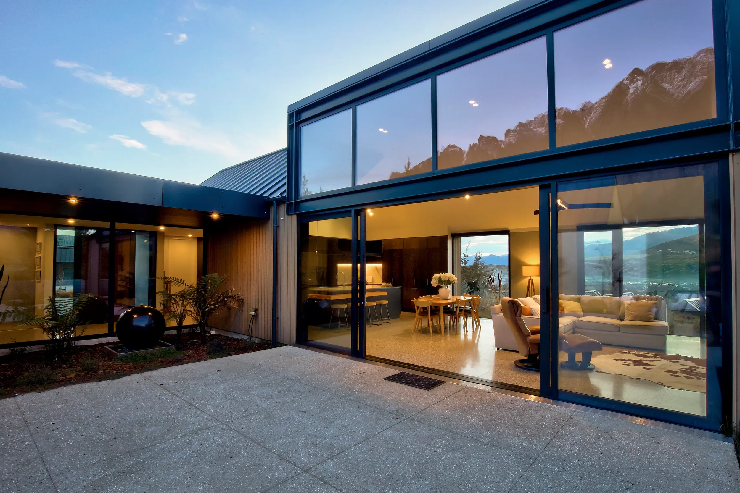 Modern house with large glass windows and sliding doors, interior living room with sofa, dining table, and kitchen, mountain view outside.