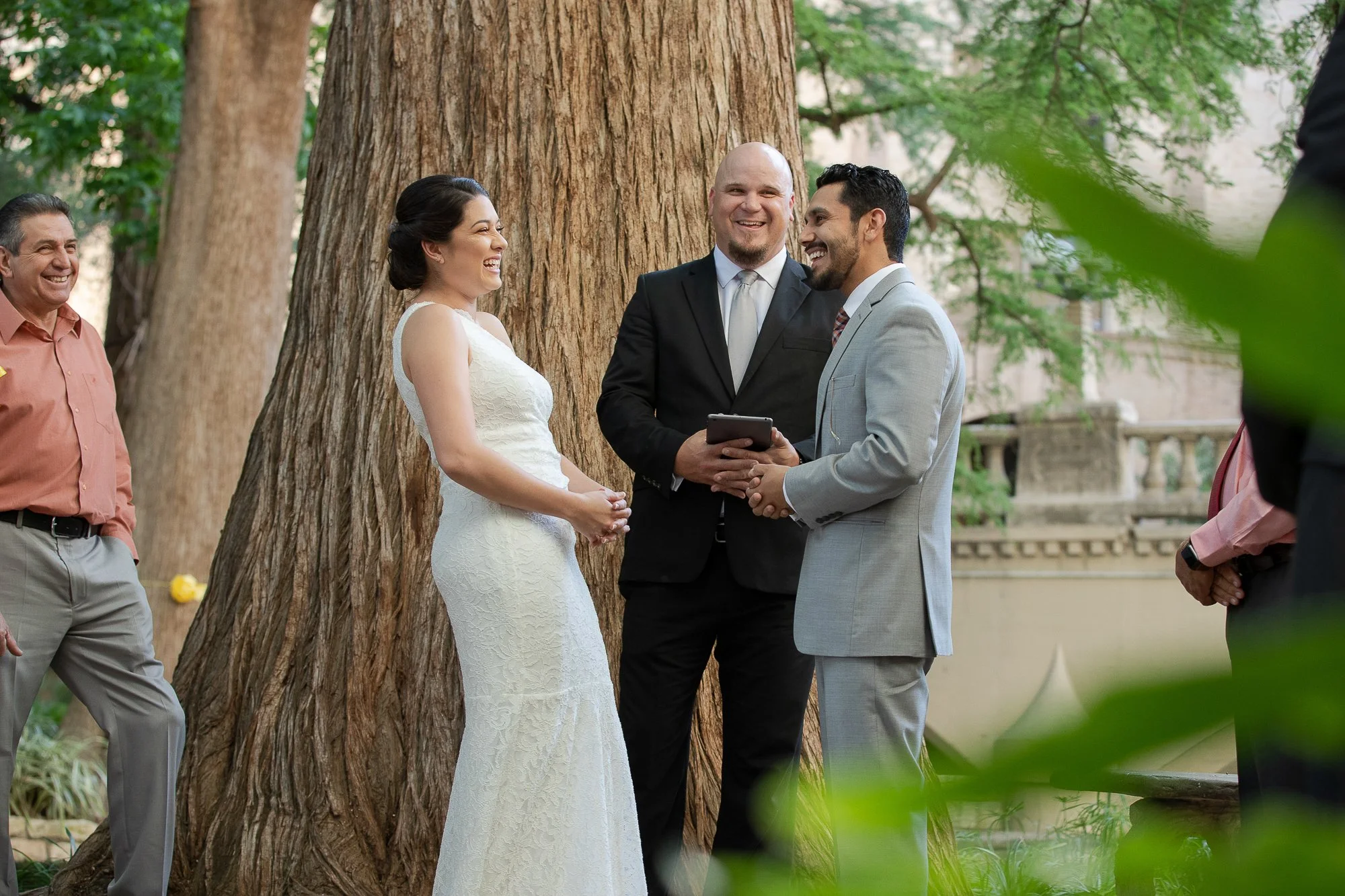 A wedding ceremony outdoors with the bride and groom holding hands and smiling at each other, surrounded by friends and family near large trees.