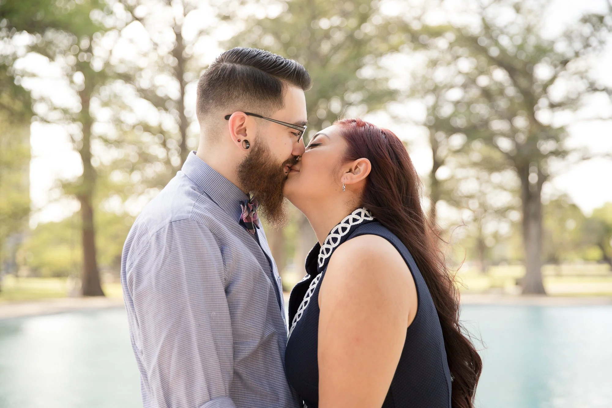 A couple kissing outdoors by a lake with trees in the background.
