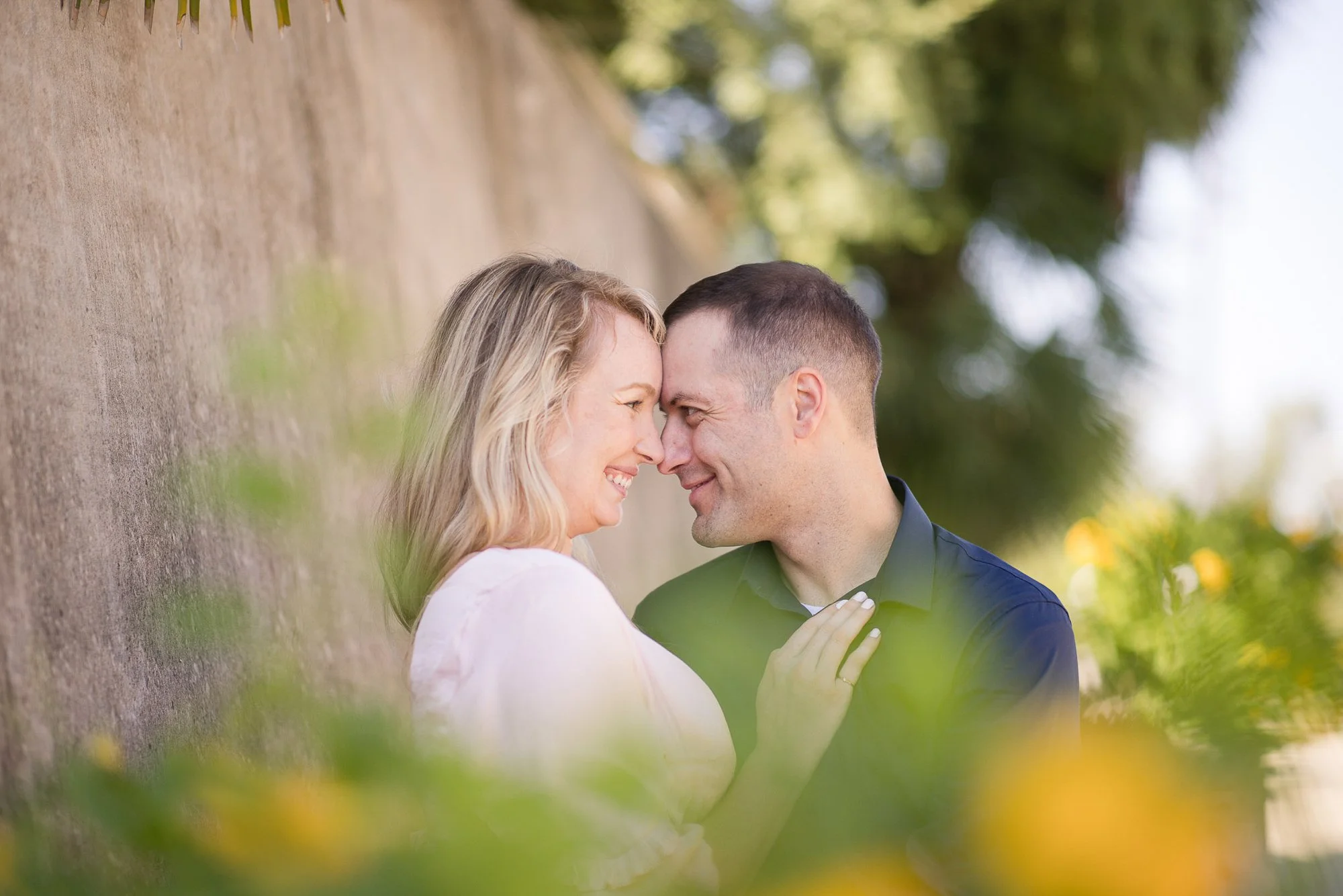 A couple with their foreheads touching and smiling at each other against a wall outdoors, with trees in the background and some blurred green and yellow foliage in the foreground.