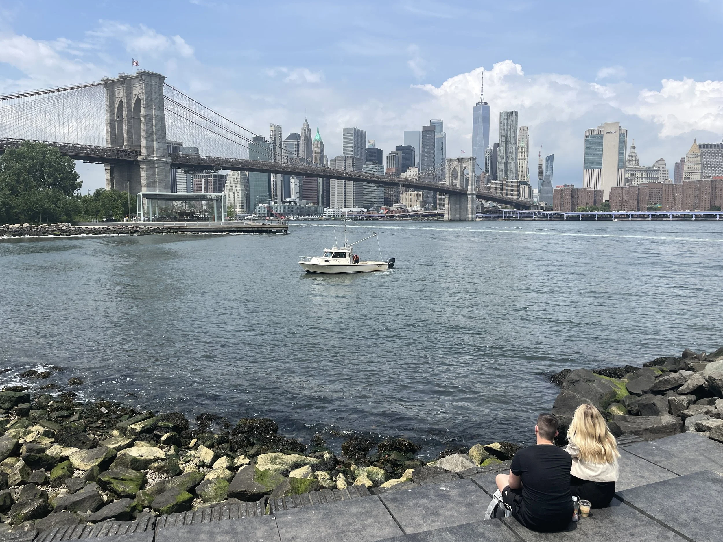 View of the New York City skyline with the Brooklyn Bridge in the foreground. Two people are sitting on the rocks by the water's edge, facing the city skyline and a boat on the water.
