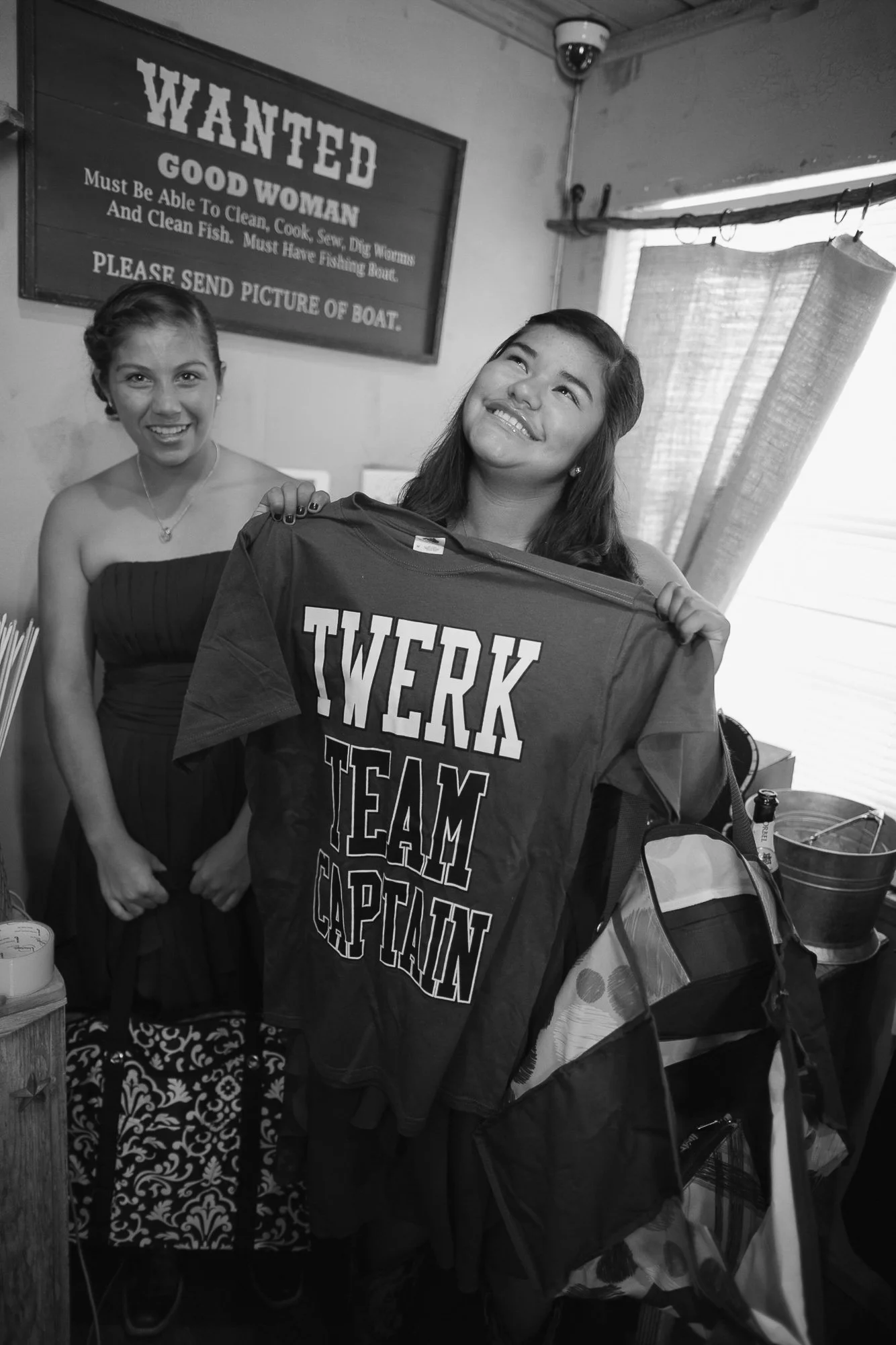 Two smiling women holding a sports jersey that reads 'TWERK TEAM CAPTAIN' inside a room with a sign that says 'WANTED GOOD WOMAN must be able to clean, cook, sew, dig worms, and clean fish. Must have fishing boat. Please send picture of boat.'