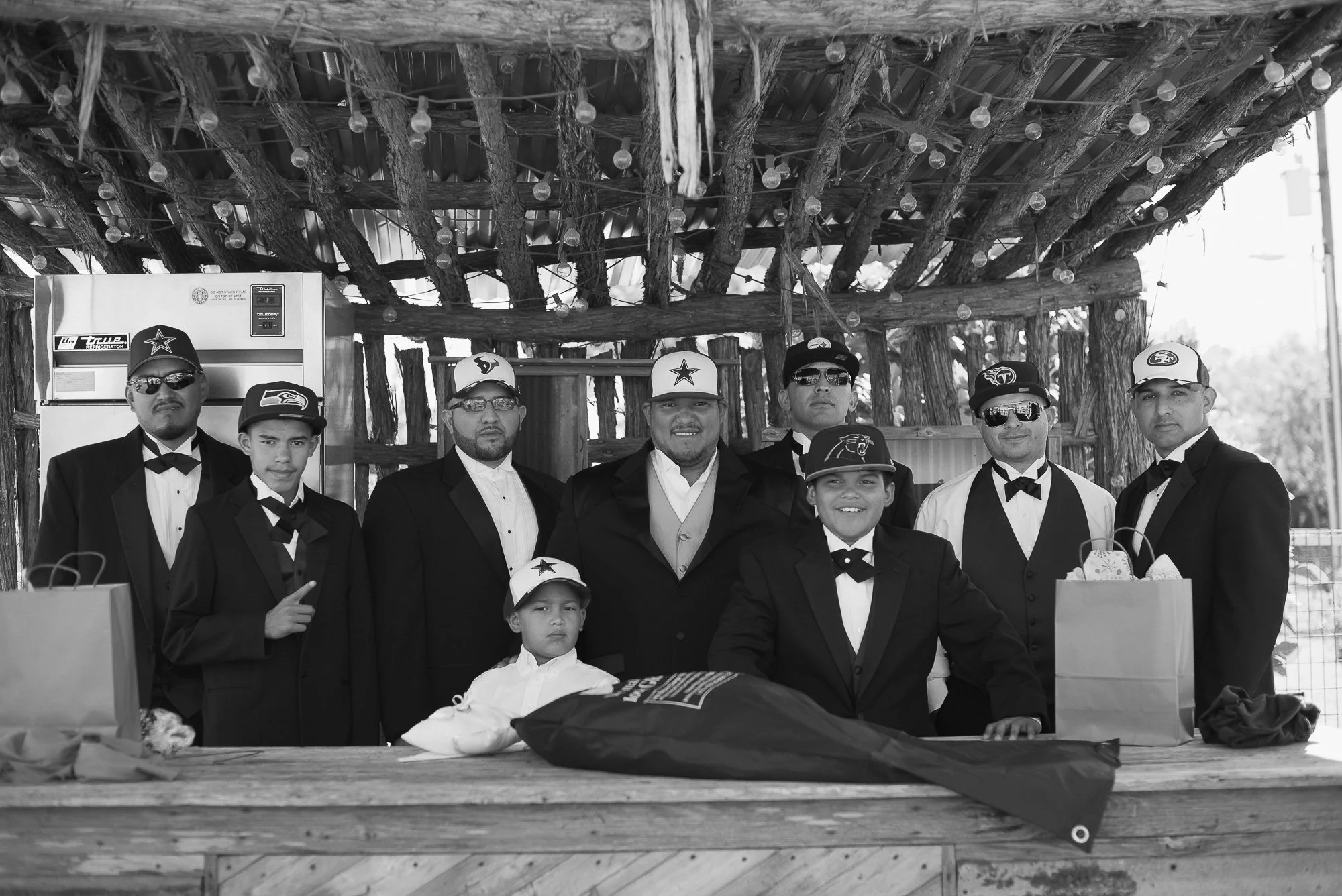 Group of men and boys in tuxedos and sport caps, standing behind a table with gifts, in front of rustic wooden structure with string lights, outdoors.