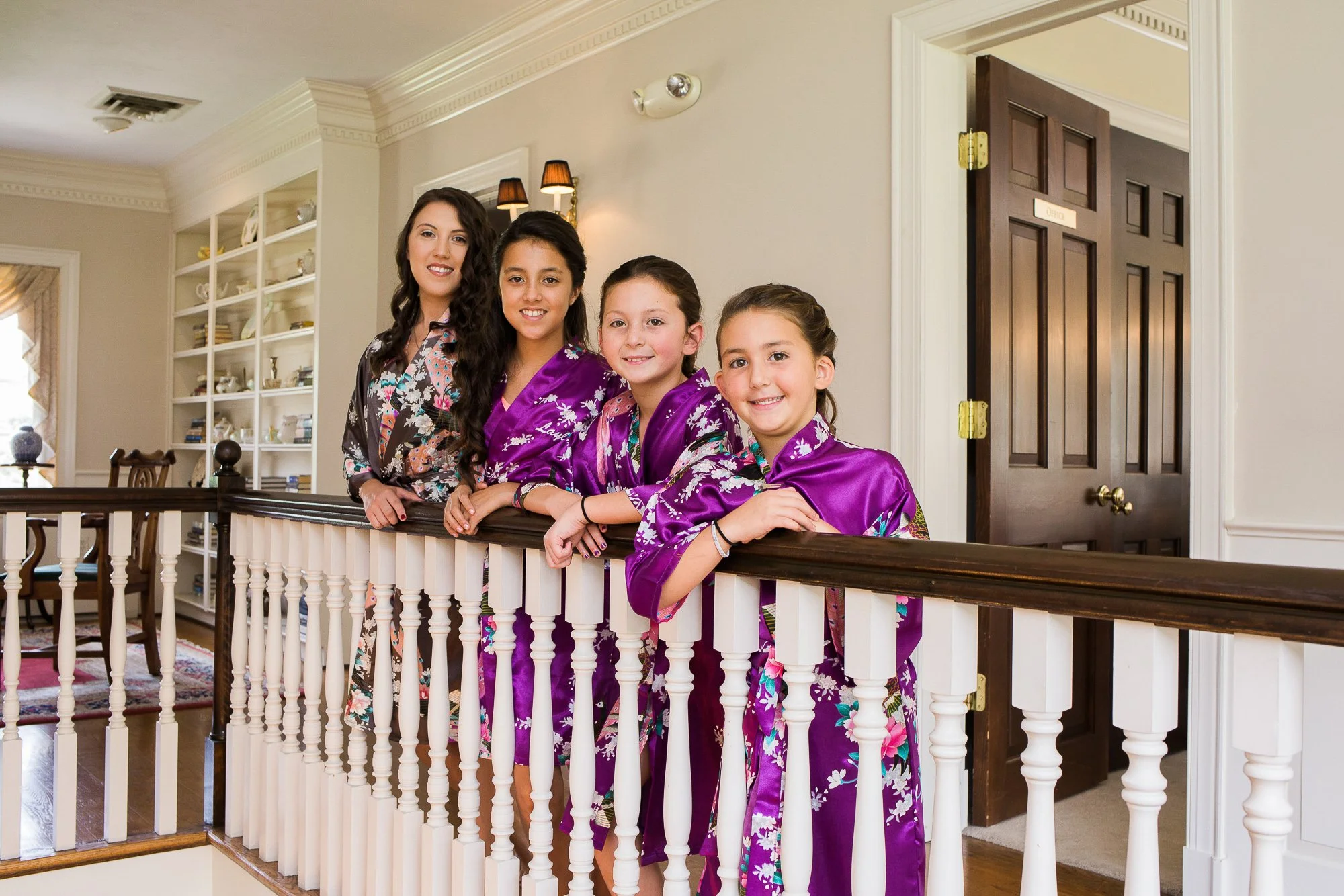 Four young girls and one woman standing on a staircase balcony inside a house. The girls are wearing purple satin robes with floral patterns, and the woman is in a floral blouse. They are smiling for the photo, with a white railing and a doorway behi