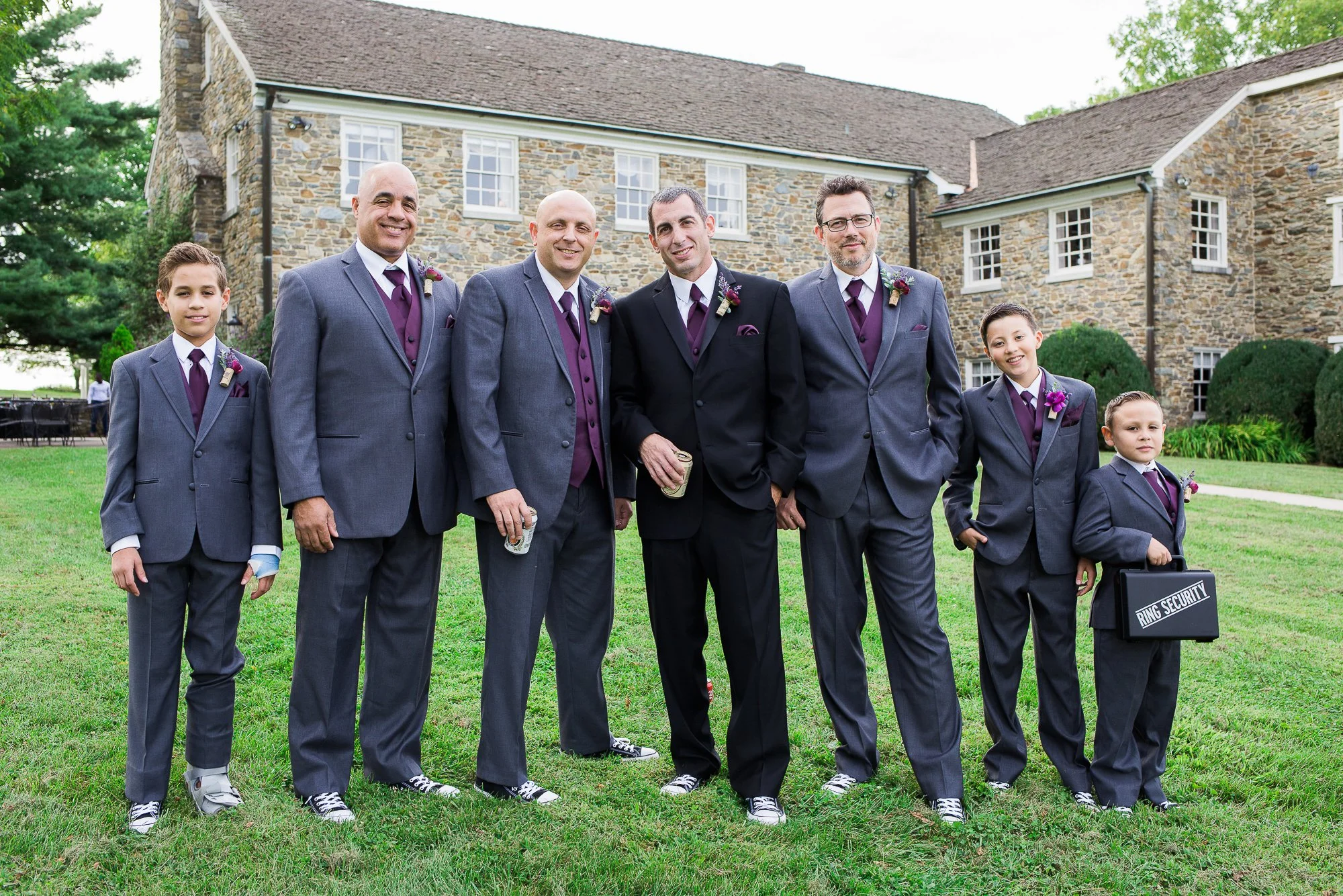 Group of eight men and boys in suits standing on grass in front of a stone building, smiling, at a wedding. Some hold drinks; one boy on the right holds a black briefcase labeled 'Ring Security'.