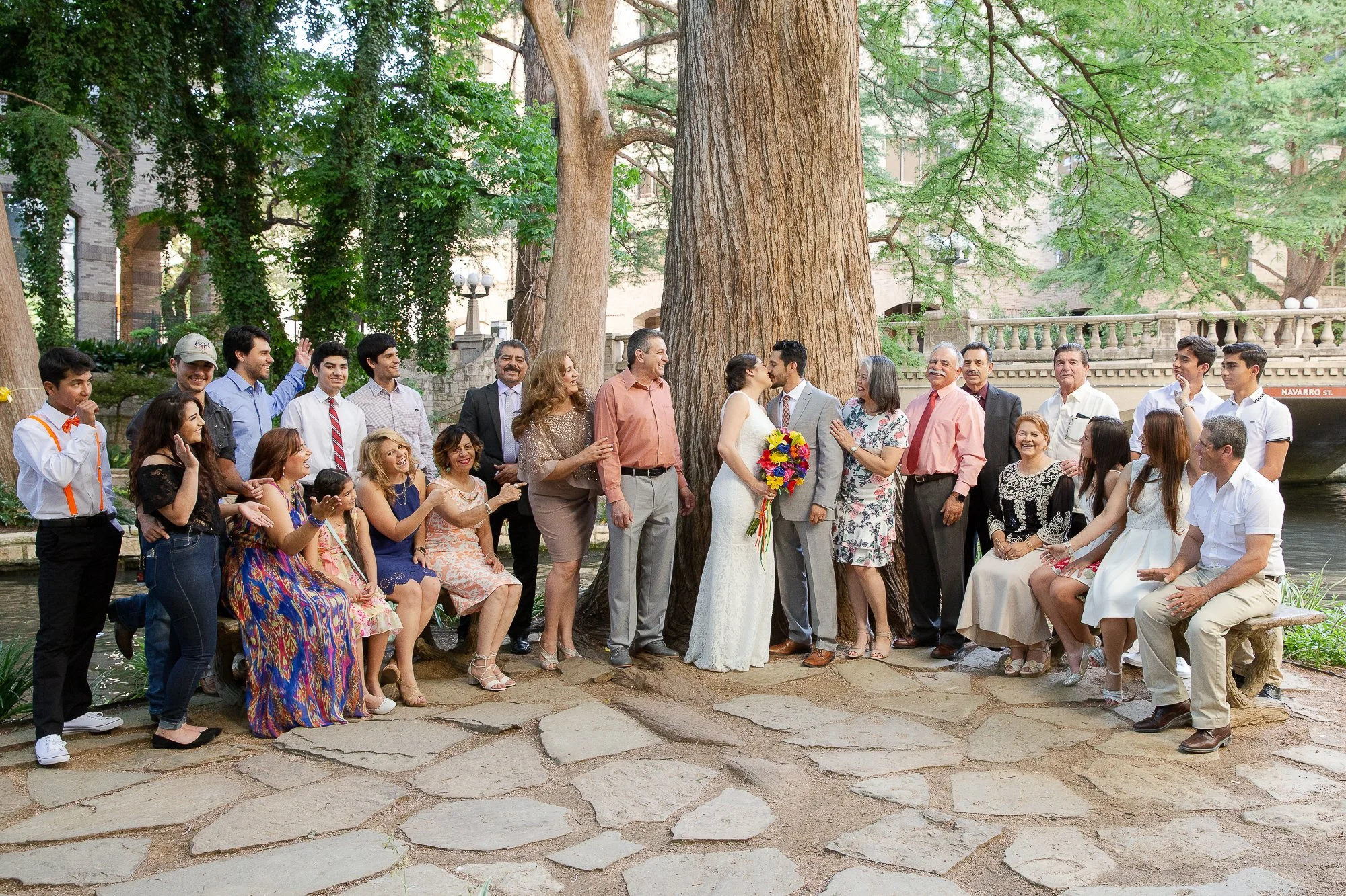 A wedding photoshoot with a bride and groom kissing in front of a large tree, surrounded by family and friends in a park setting.