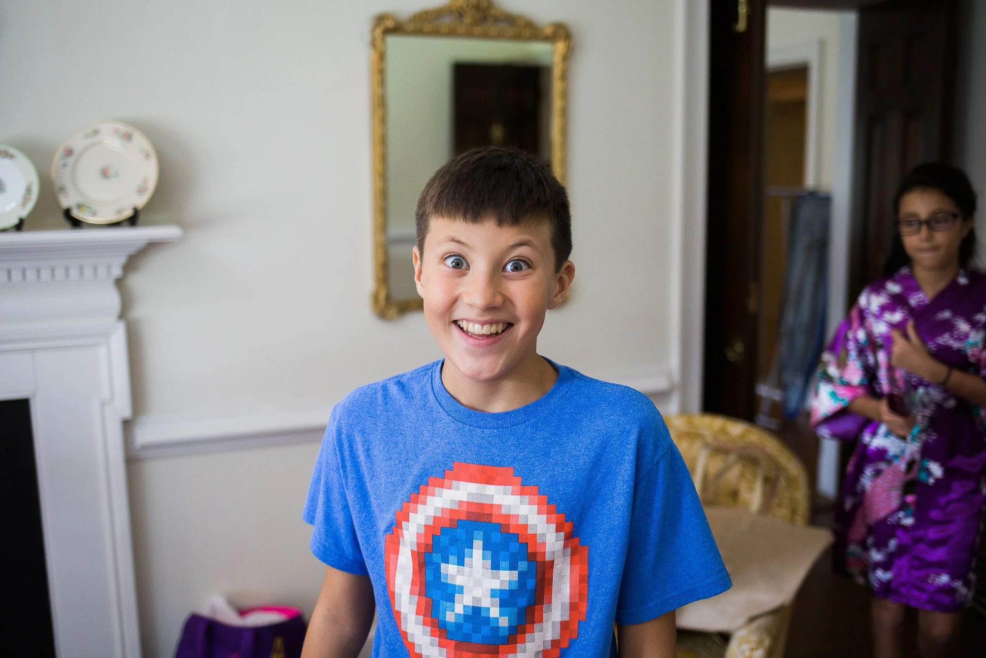 A young boy with short brown hair, wide eyes, and a big smile, wearing a blue T-shirt with a pixelated Captain America shield design, standing in a room with a woman in a purple kimono in the background.