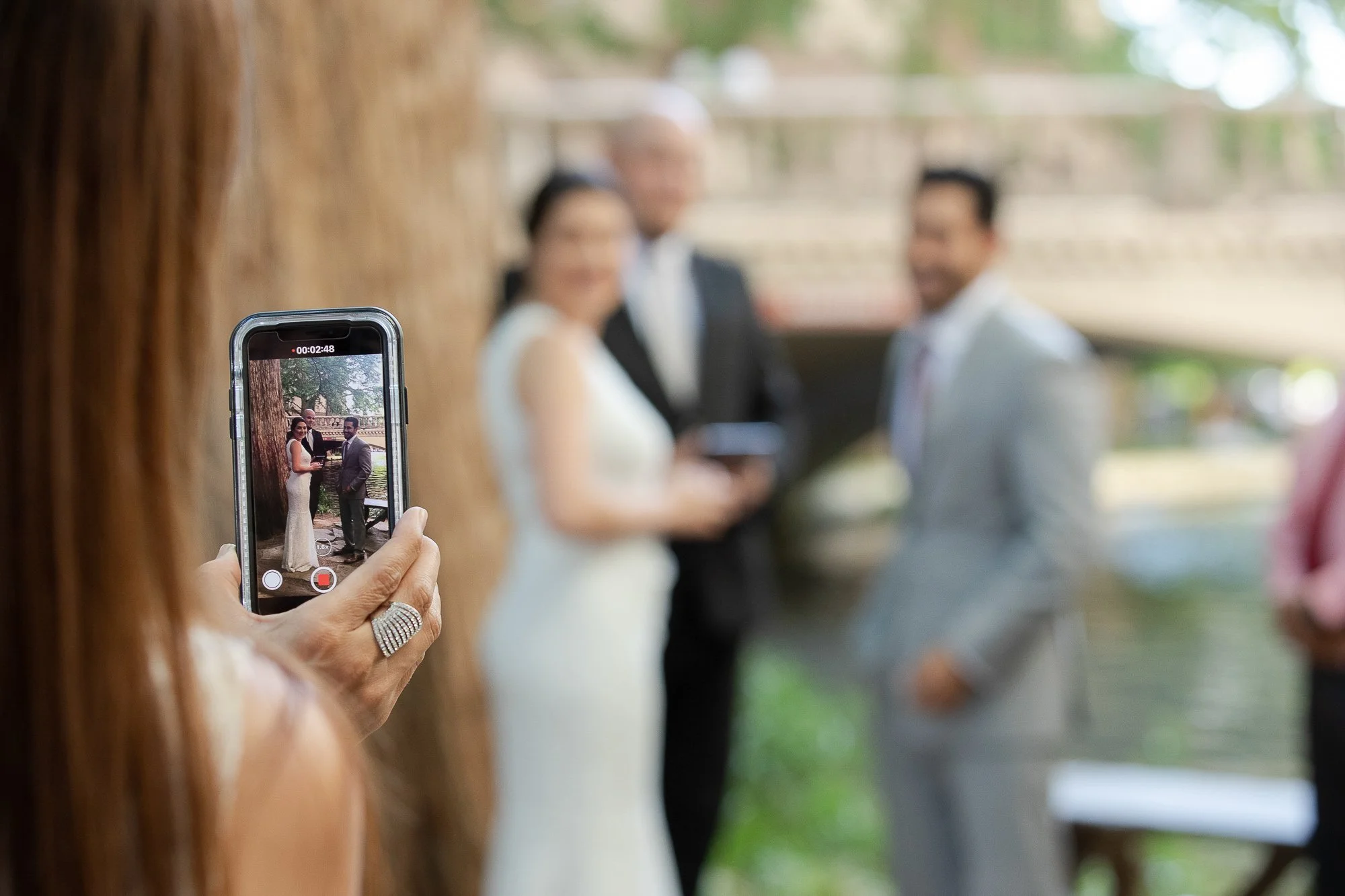 Person taking a photo of a wedding ceremony with a smartphone, with the bride and groom in focus on the phone's screen