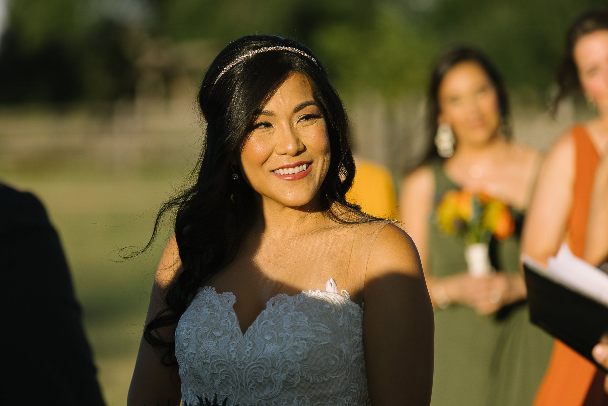 A woman with long dark hair and a sparkling headband smiling at an outdoor event, with other women and a man in the background, holding flowers and papers.