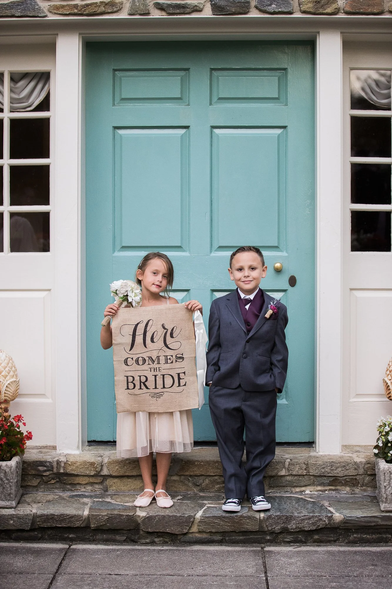 A young girl and boy standing in front of a house with a teal door. The girl is holding a bouquet and a sign that says "Here Comes the Bride". The boy is dressed in a tuxedo.