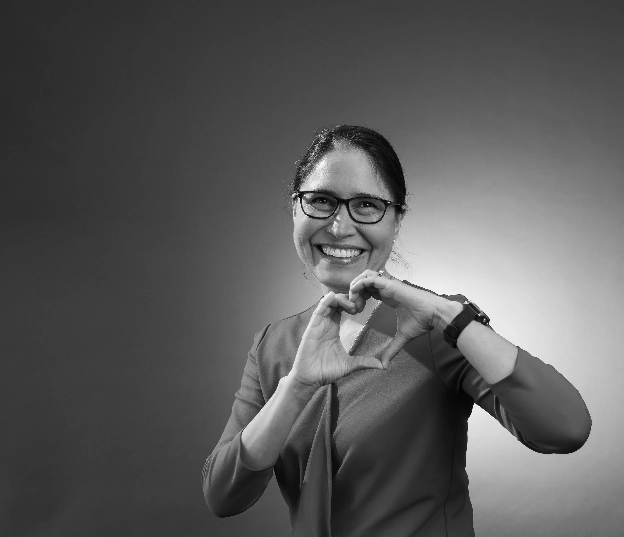 A smiling woman wearing glasses making a heart shape with her hands, standing against a plain background.