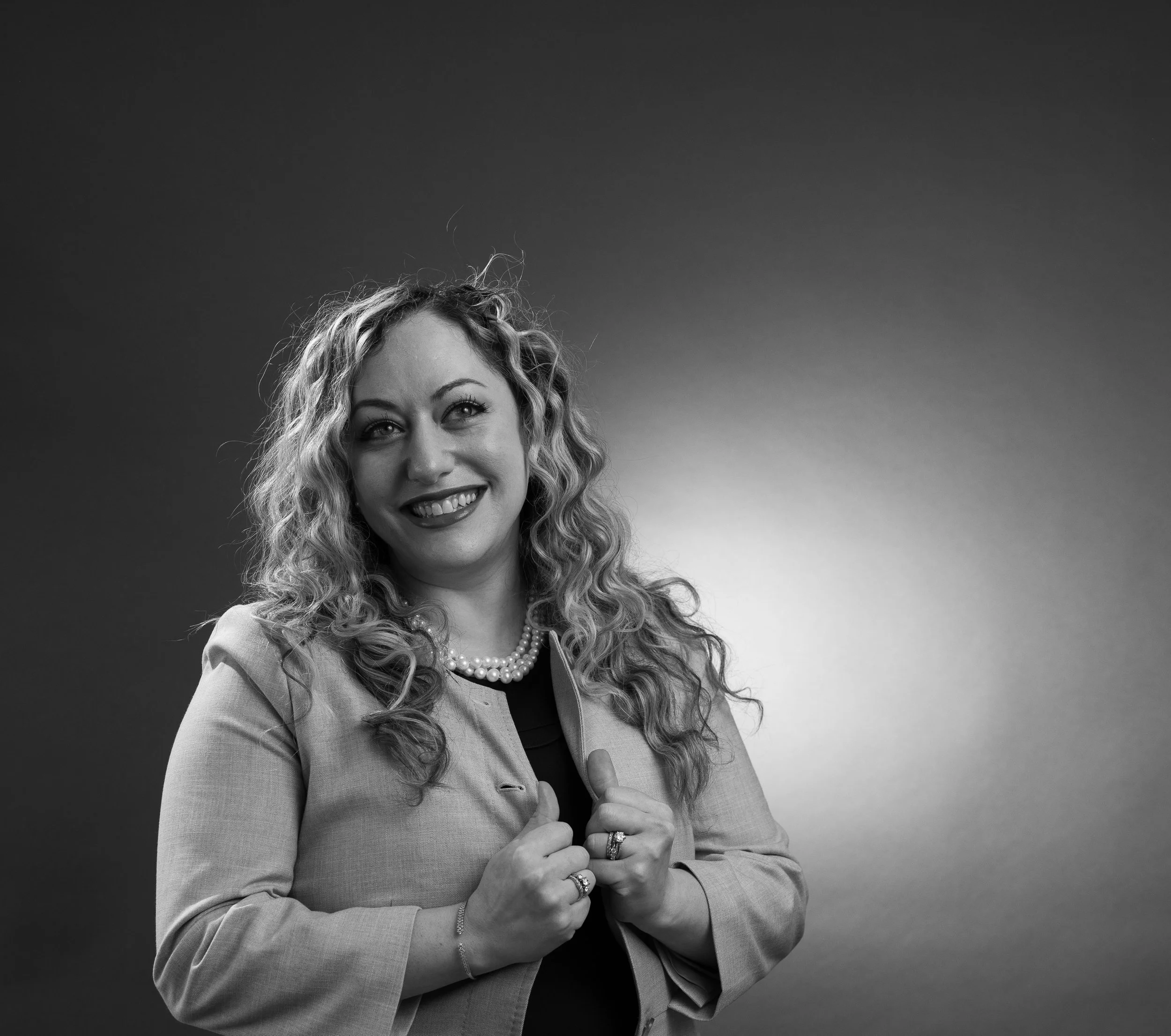 Black and white portrait of a woman with curly hair, smiling, wearing a blazer, pearl necklace, and rings, holding her blazer lapels with both hands.