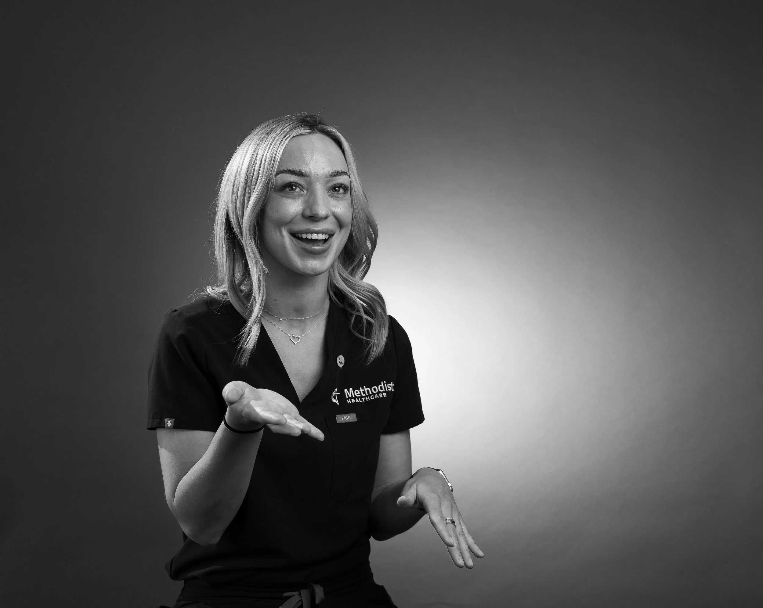 A young woman with long blonde hair, smiling and gesturing with her hands, wearing a dark medical uniform with 'Methodist Healthcare' embroidered on it and a name tag, standing against a plain gradient background.