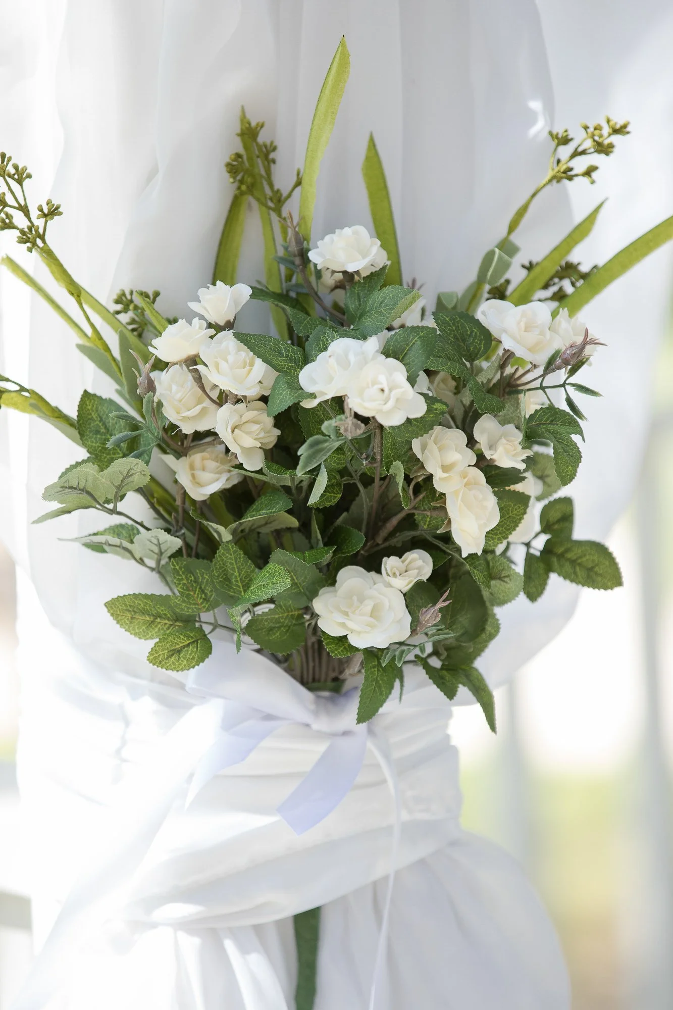 A bouquet of white flowers and green leaves wrapped with white fabric and a white ribbon, placed near a window with sunlight.