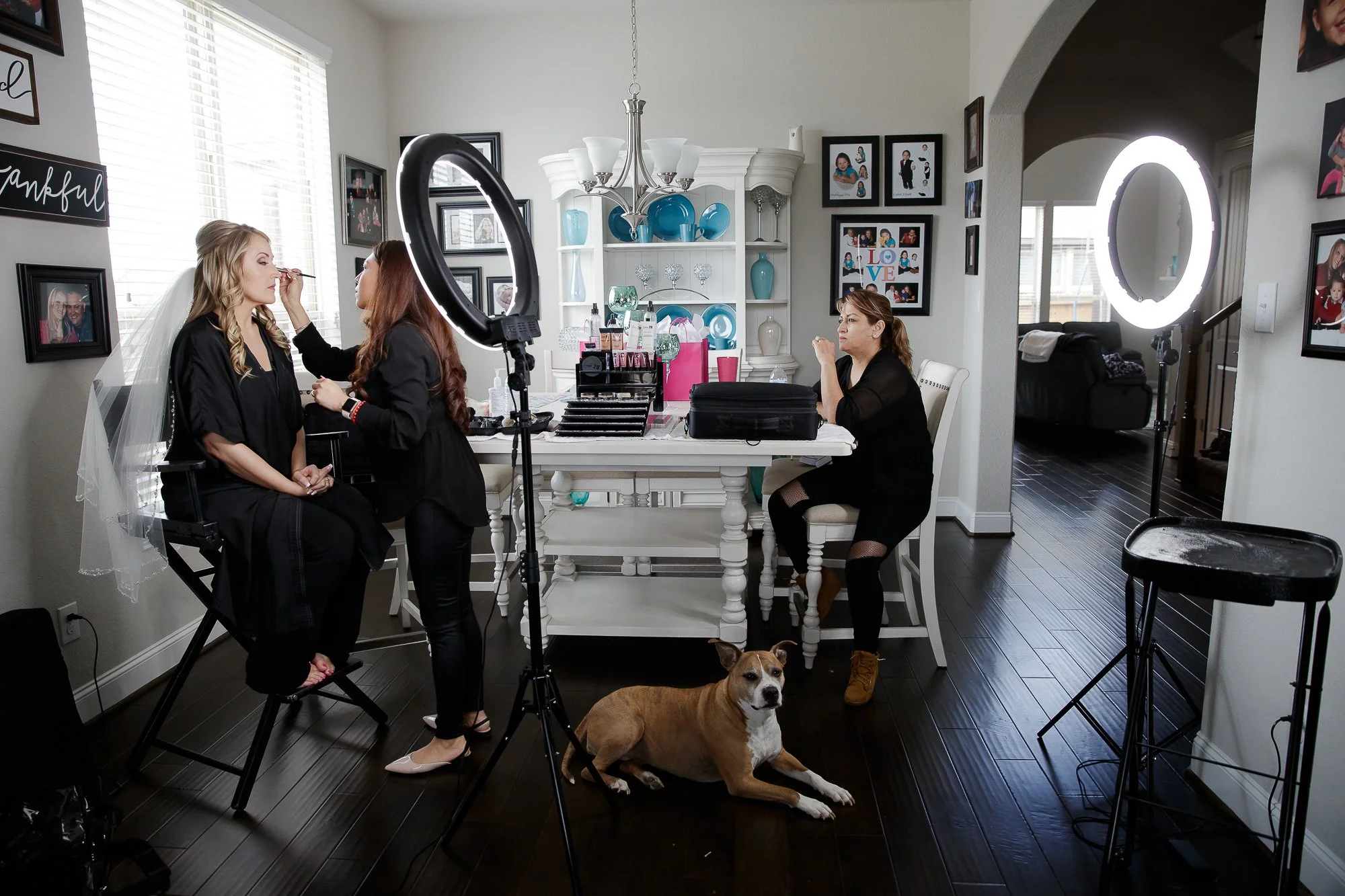 A woman has her makeup done by a makeup artist while another woman is seated at a table. There are photography lighting equipment and a dog lying on the dark wooden floor in a well-lit dining room with picture frames and decorative items on the walls
