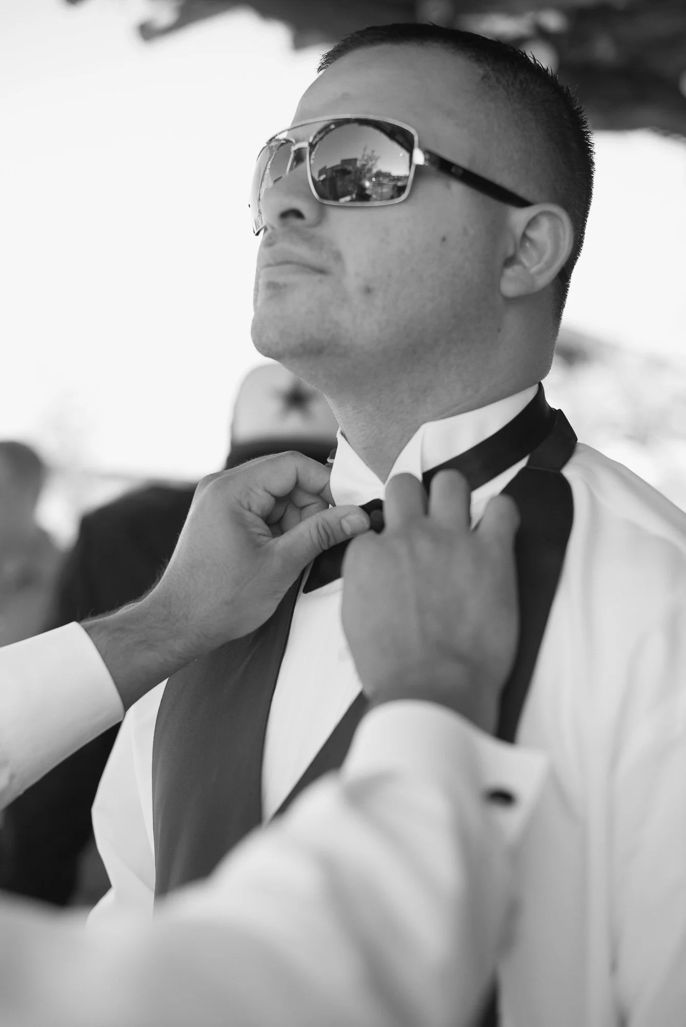 A man wearing sunglasses and formal attire is having a bow tie adjusted by another person, likely during a formal event or celebration.