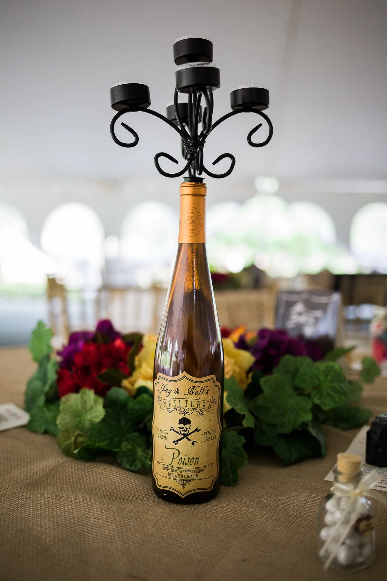 Decorative bottle with a candle holder on top, featuring a Halloween-themed label with skull and crossbones, labeled 'Poison' and 'Unfiltered', placed on a table with floral decorations.