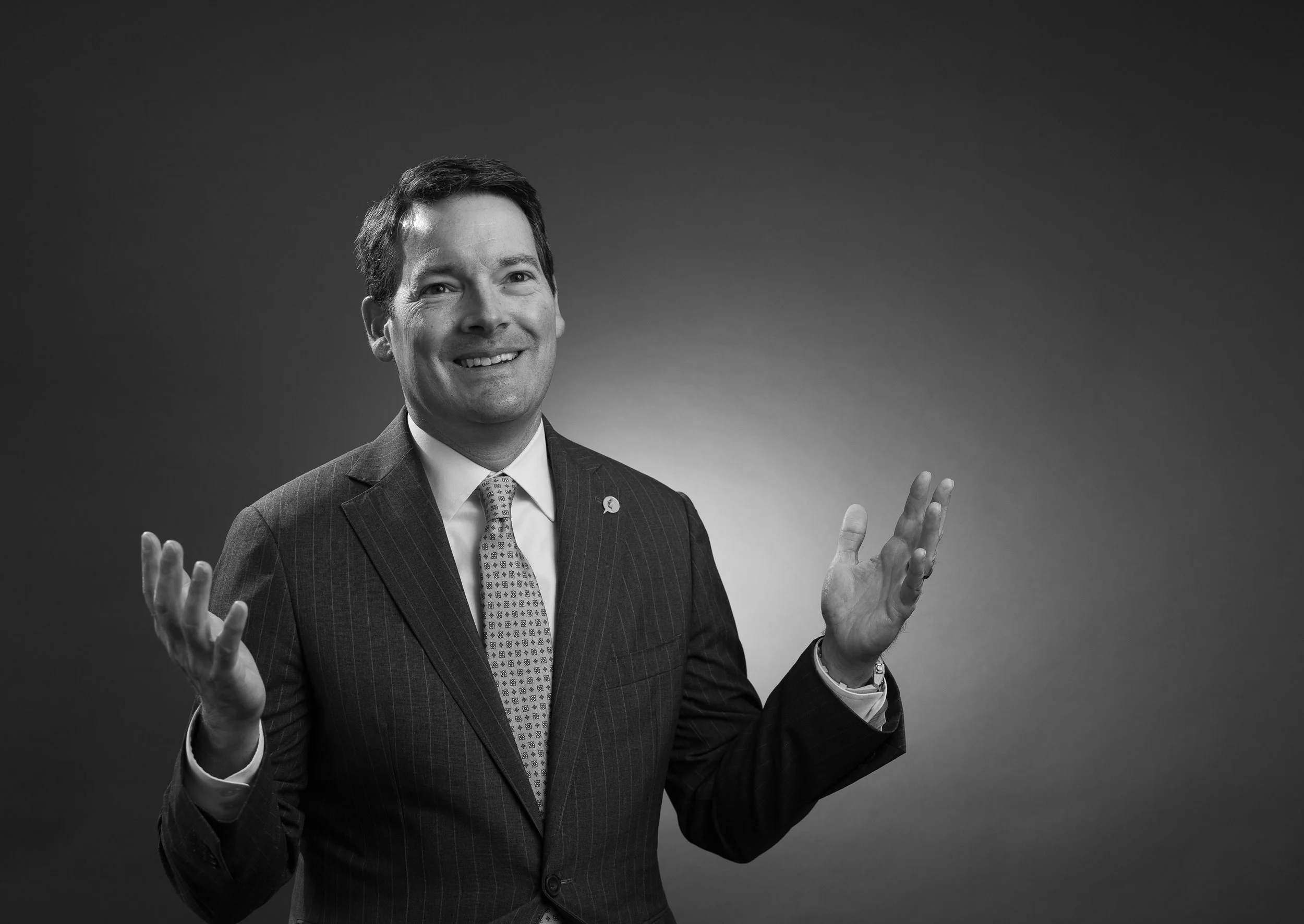 A man in a suit smiling, with his hands raised in a gesturing manner, against a plain background.