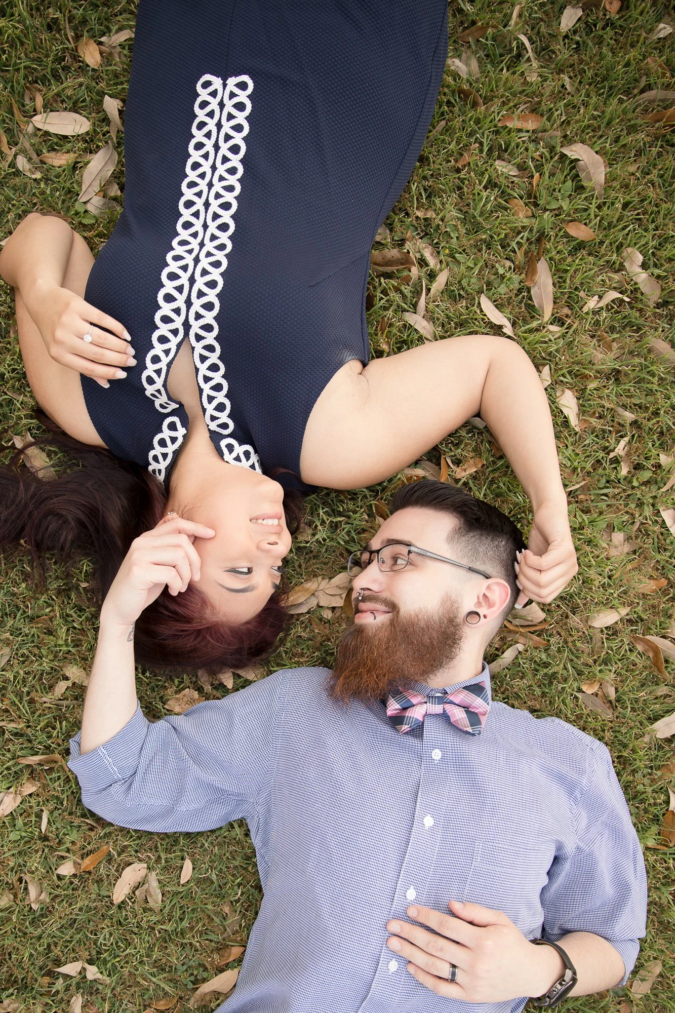 A man with glasses and a groomed beard lying on the grass, looking at a woman with dark hair and a ring on her finger, also lying on the grass. They are smiling and looking at each other.