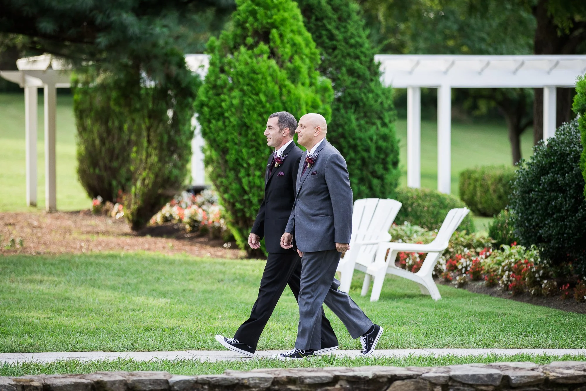 Two men in suits walking outdoors in a garden with green bushes and flowers, one of them trying on sneakers.