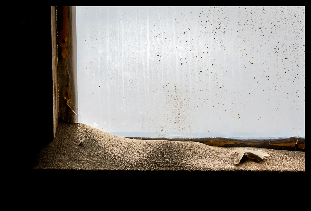 Condensation lines of water on a window pane run down into the sand that has built up on the window-sill.