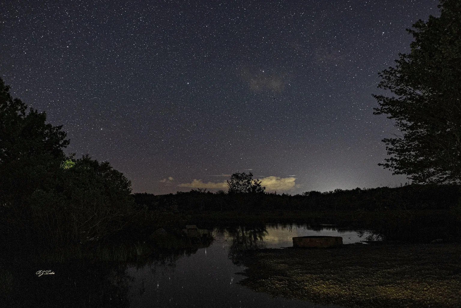 Astronomical twilight has just ended and true darkness is now beginning at around 11:24 PM, one day after summer solstice. Light pollution in the background emanates from a community to the west of here. It might well be from Yarmouth. Light has a very long reach. The photo was taken beside the Quinan River, down at the firepit area. Heavy rains have flooded this spot so the firepit is underwater and the furniture has been relocated to drier ground. This, too, shall pass.