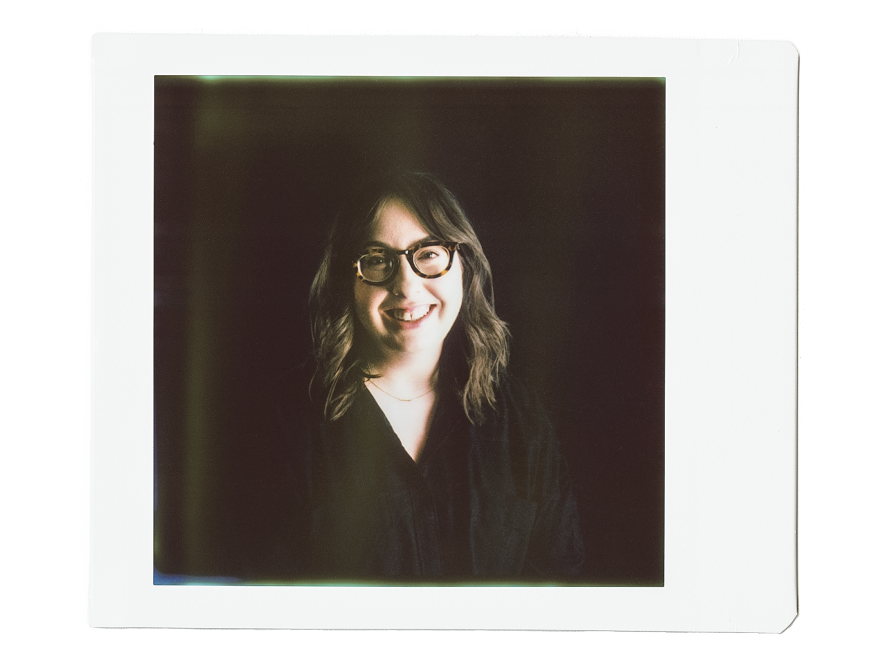 Close-up of a young woman with shoulder-length wavy brown hair, wearing glasses, smiling, against a dark background, framed by a blurred white border.