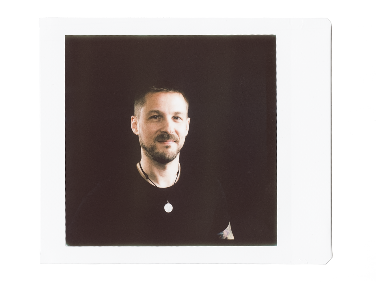 A young man with short brown hair and a beard, wearing a black shirt and a necklace, posing for a portrait against a dark background.