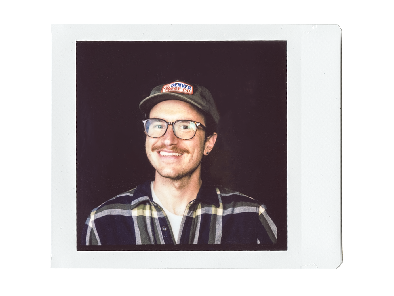 A man with glasses, a mustache, and a beard wears a black cap with a 'Denver Beer Co' logo and a plaid shirt. He is smiling and facing slightly to the left against a dark background.