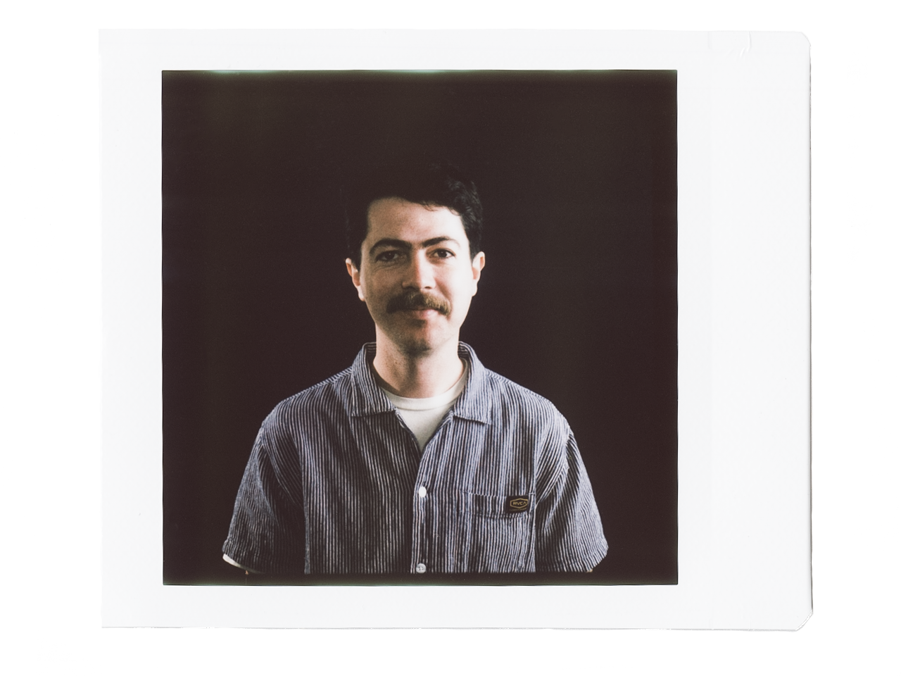 A man with dark hair and facial hair, wearing a striped button-up shirt, looks at the camera with a neutral expression against a dark background.
