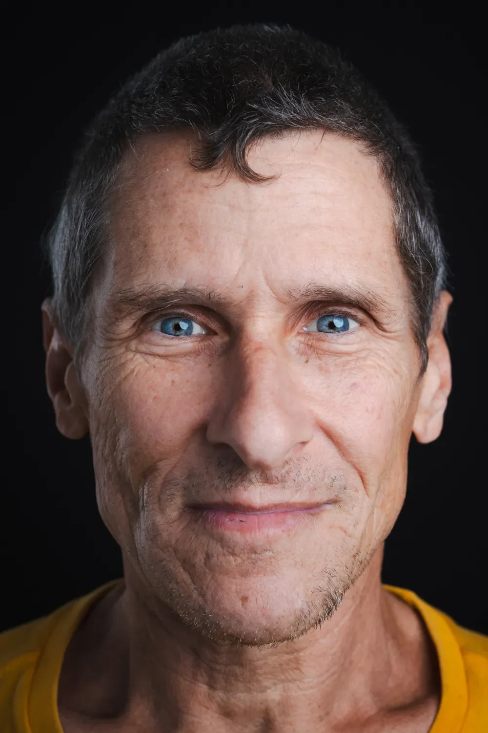Close-up portrait of a middle-aged man with blue eyes, short dark hair with gray streaks, a light facial stubble, and wearing a yellow shirt, against a black background.