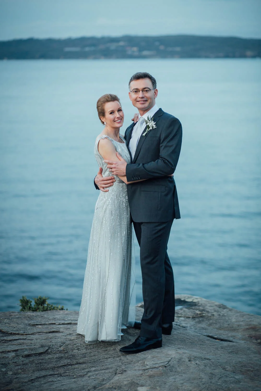 Couple in formal attire posing on a rocky shoreline with water in the background.