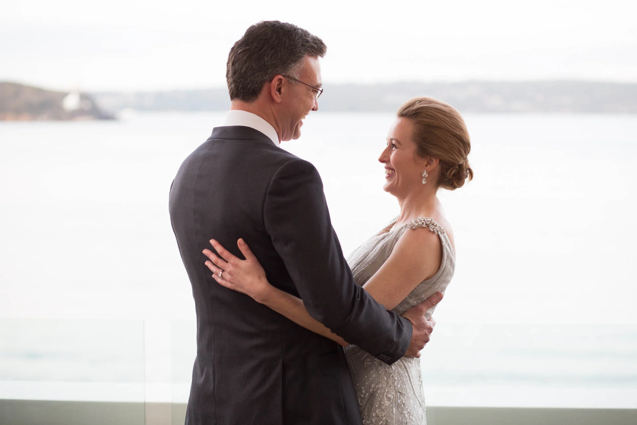 A couple in formal attire embracing and smiling near a scenic waterfront view.