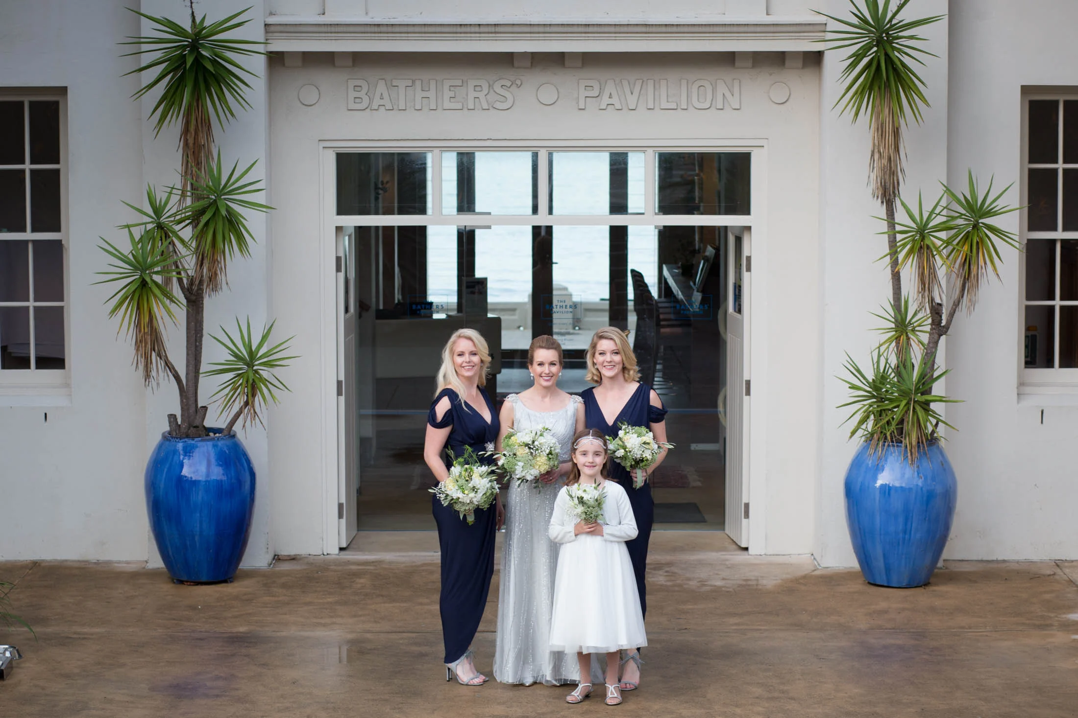 Four women in formal attire holding bouquets outside Bathers' Pavilion entrance with blue planters.