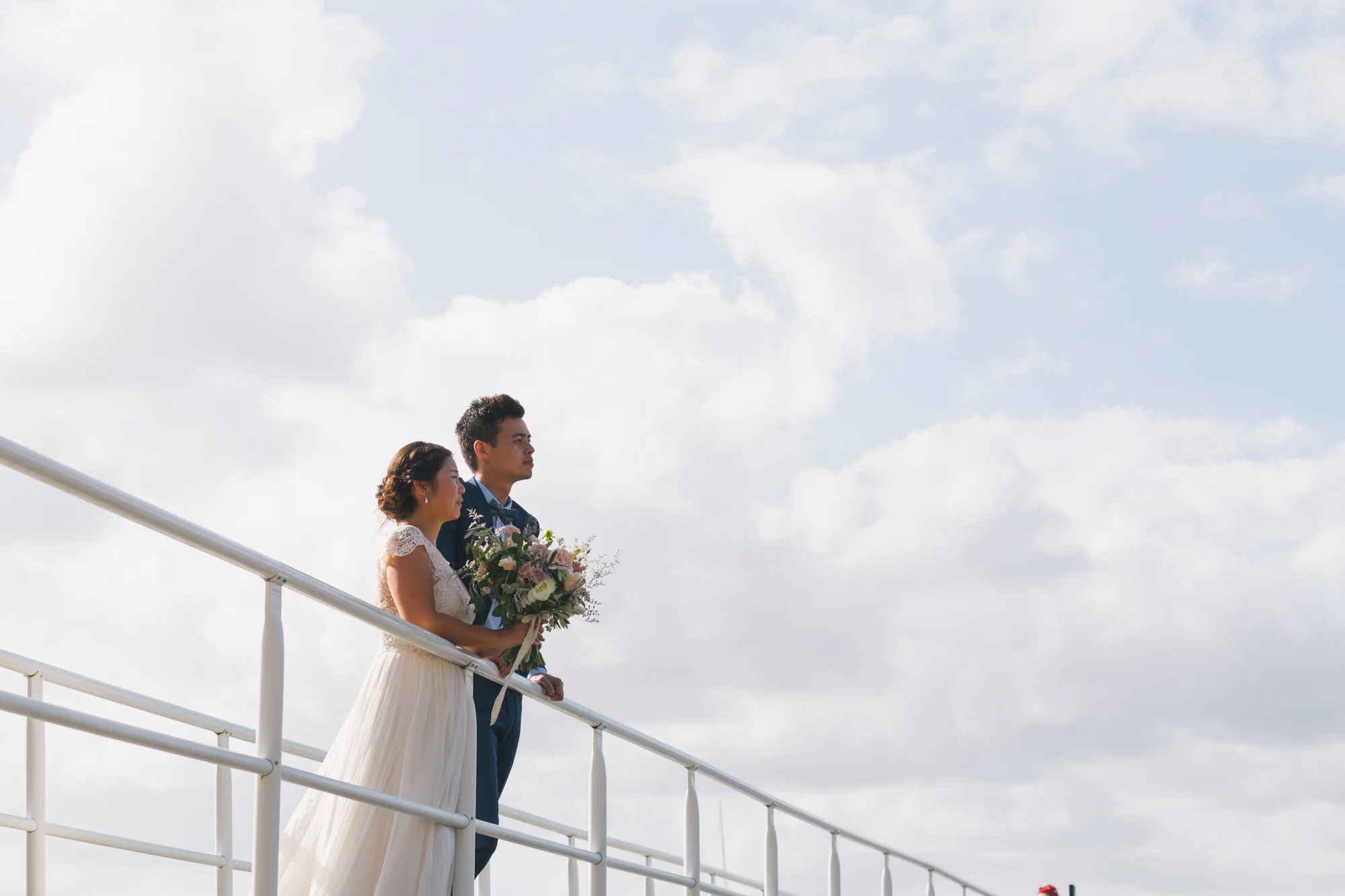 A bride and groom standing on a boat deck, holding a bouquet of flowers, with a blue sky and white clouds in the background.