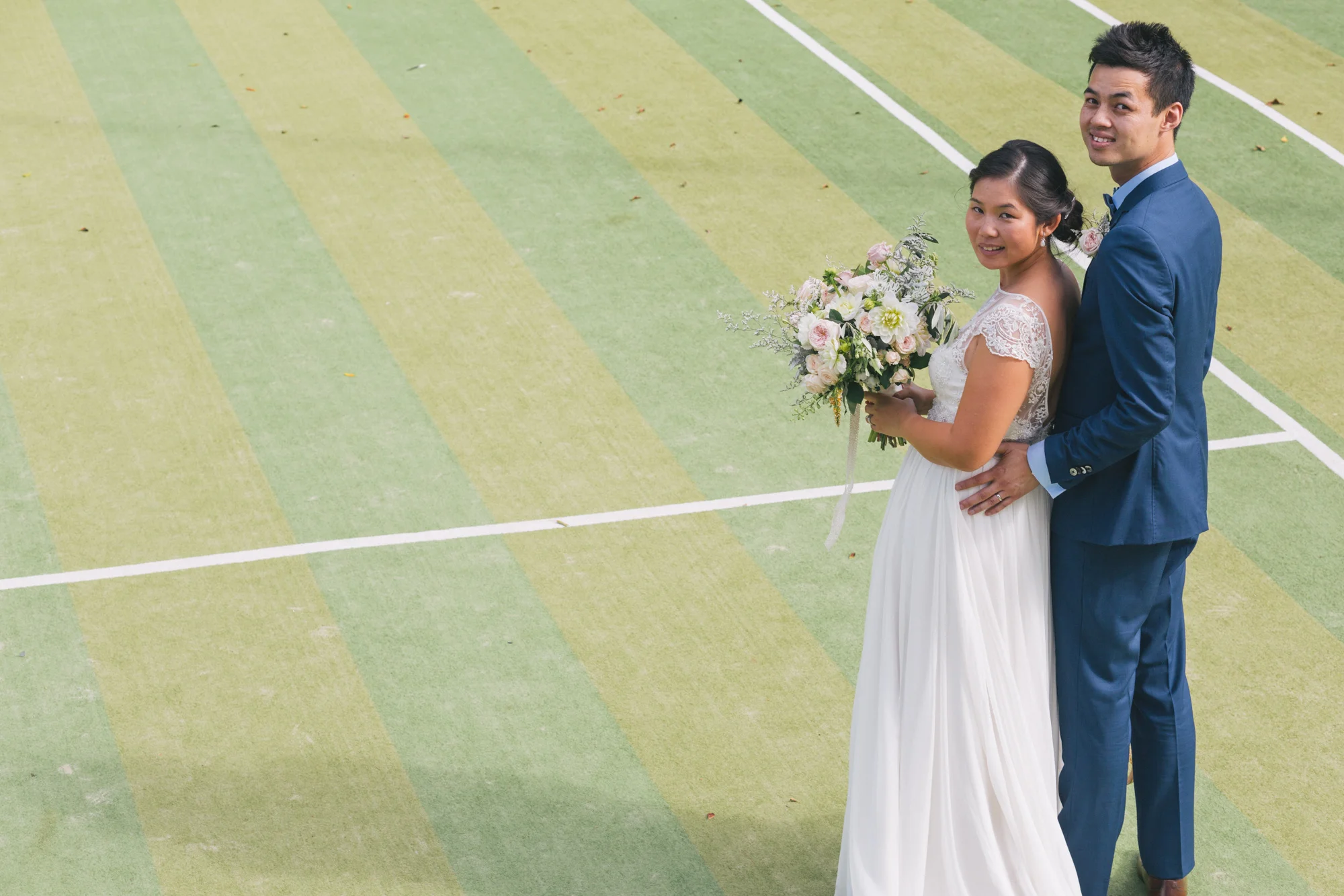A wedding couple standing on a green sport court, with the bride holding a bouquet of flowers and the groom in a blue suit with a bow tie, both smiling.