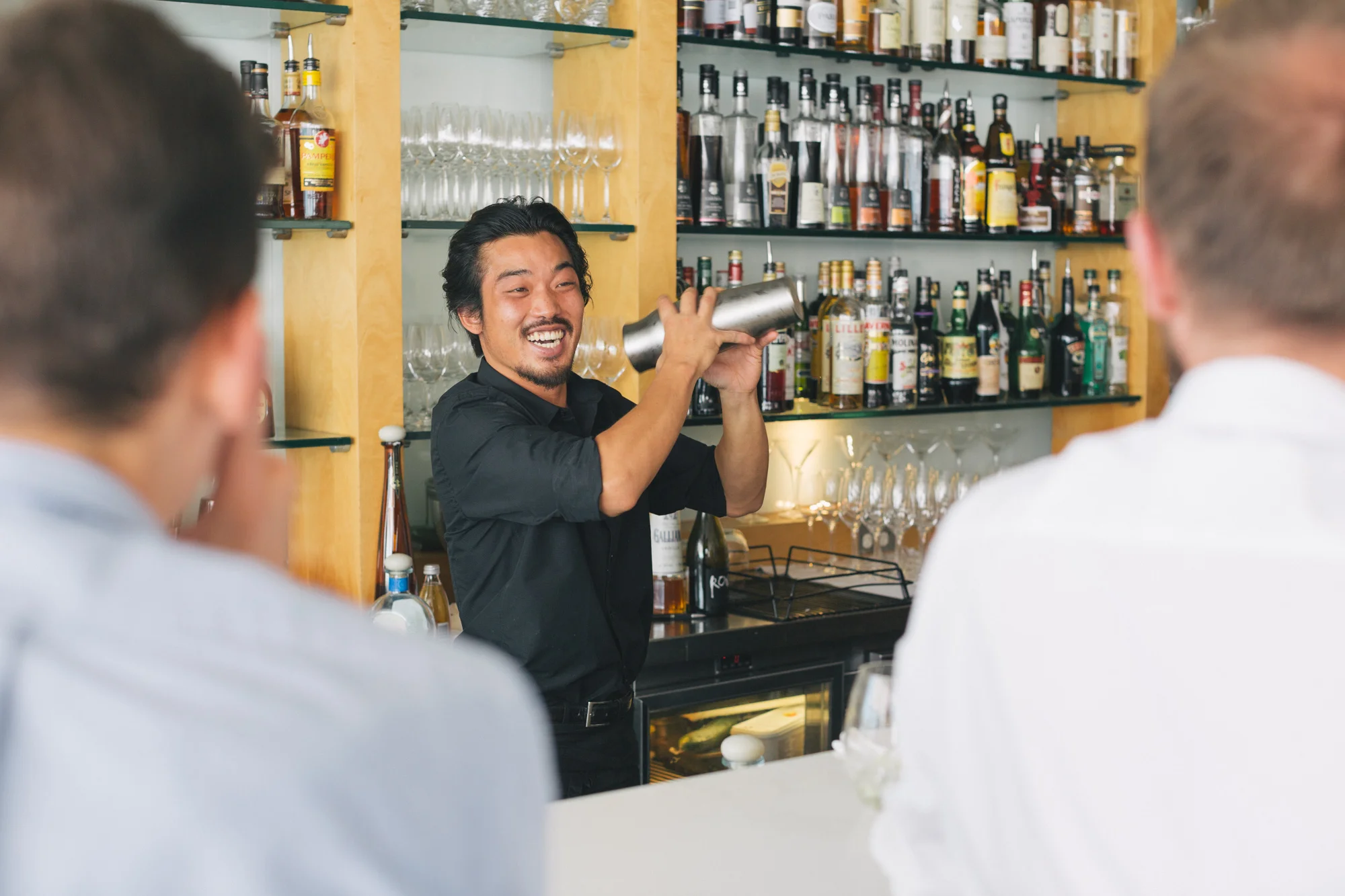A bartender with black hair and a black shirt smiling and pouring a drink for customers at a bar with shelves of liquor bottles and glasses in the background.