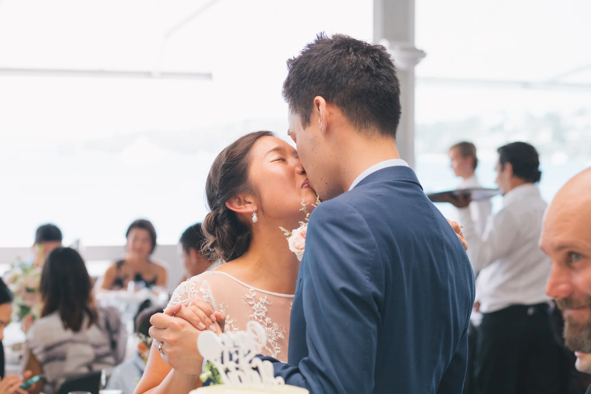 A bride and groom share a kiss during their wedding reception, surrounded by guests in a brightly lit indoor venue.