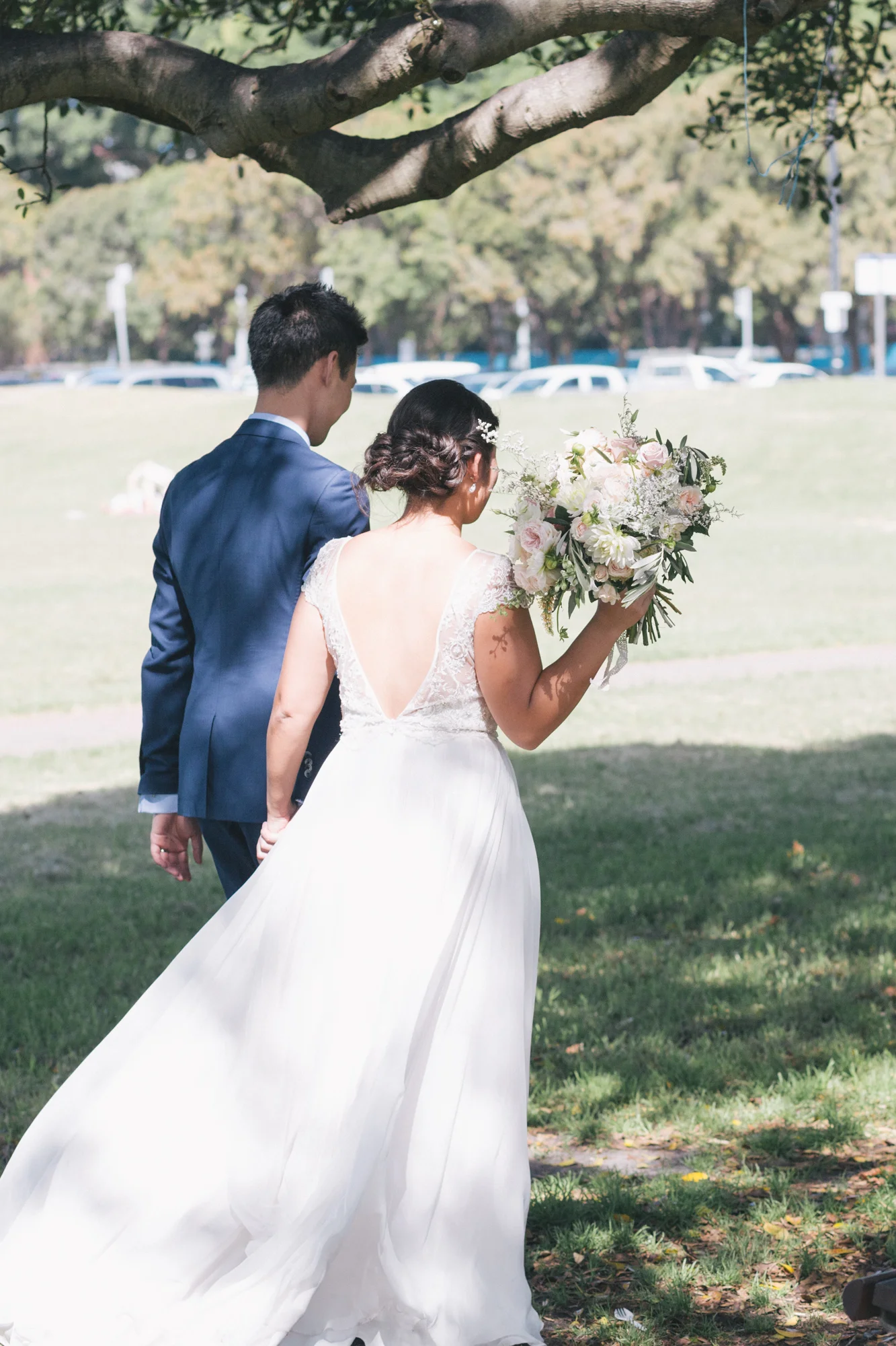 Bride and groom walking hand in hand, bride holding a bouquet of flowers, outdoor setting with greenery.