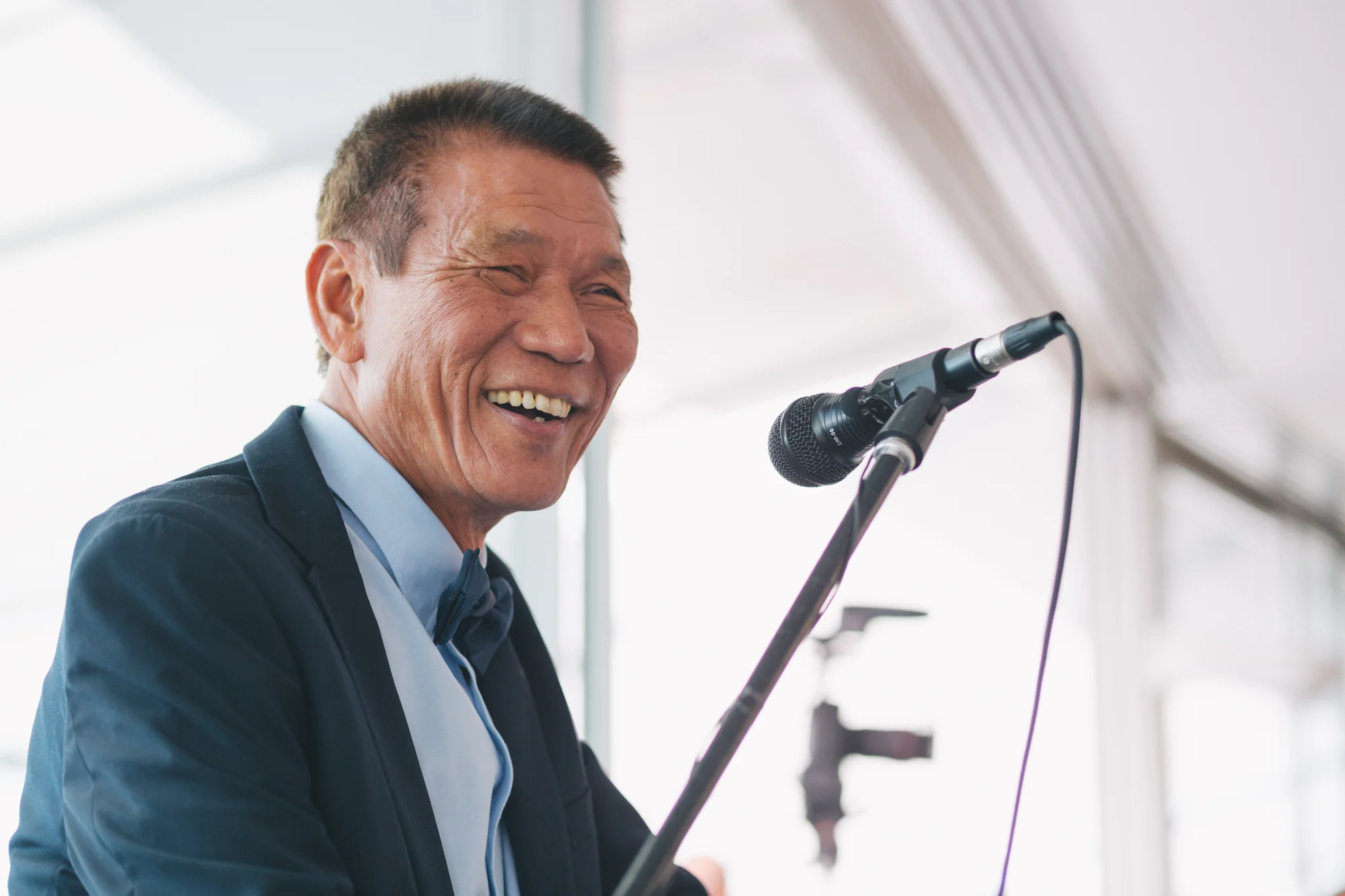 An elderly man in formal attire, including a dark suit and a bow tie, is smiling while speaking or singing into a microphone at an event or celebration.