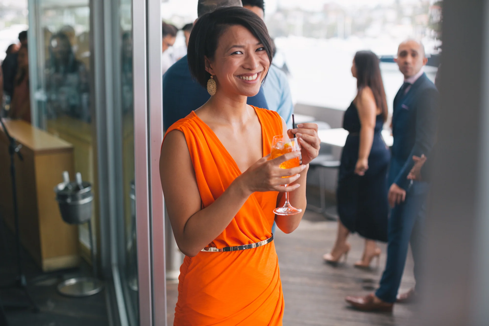 A woman in an orange dress holding a glass of orange cocktail at a social event, smiling at the camera, with people in formal attire in the background.