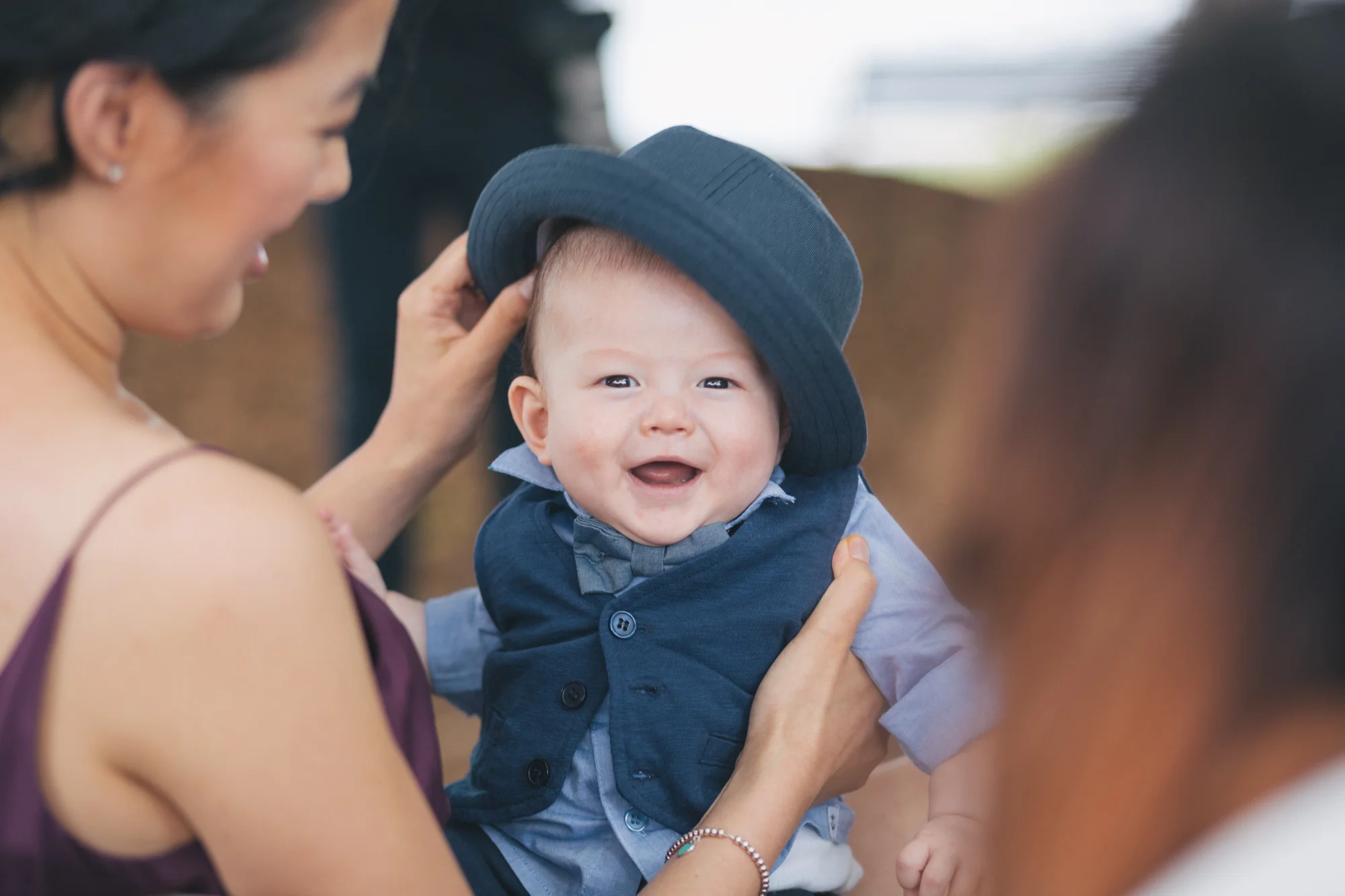 A woman is holding a smiling baby boy wearing a blue outfit and a dark blue hat.